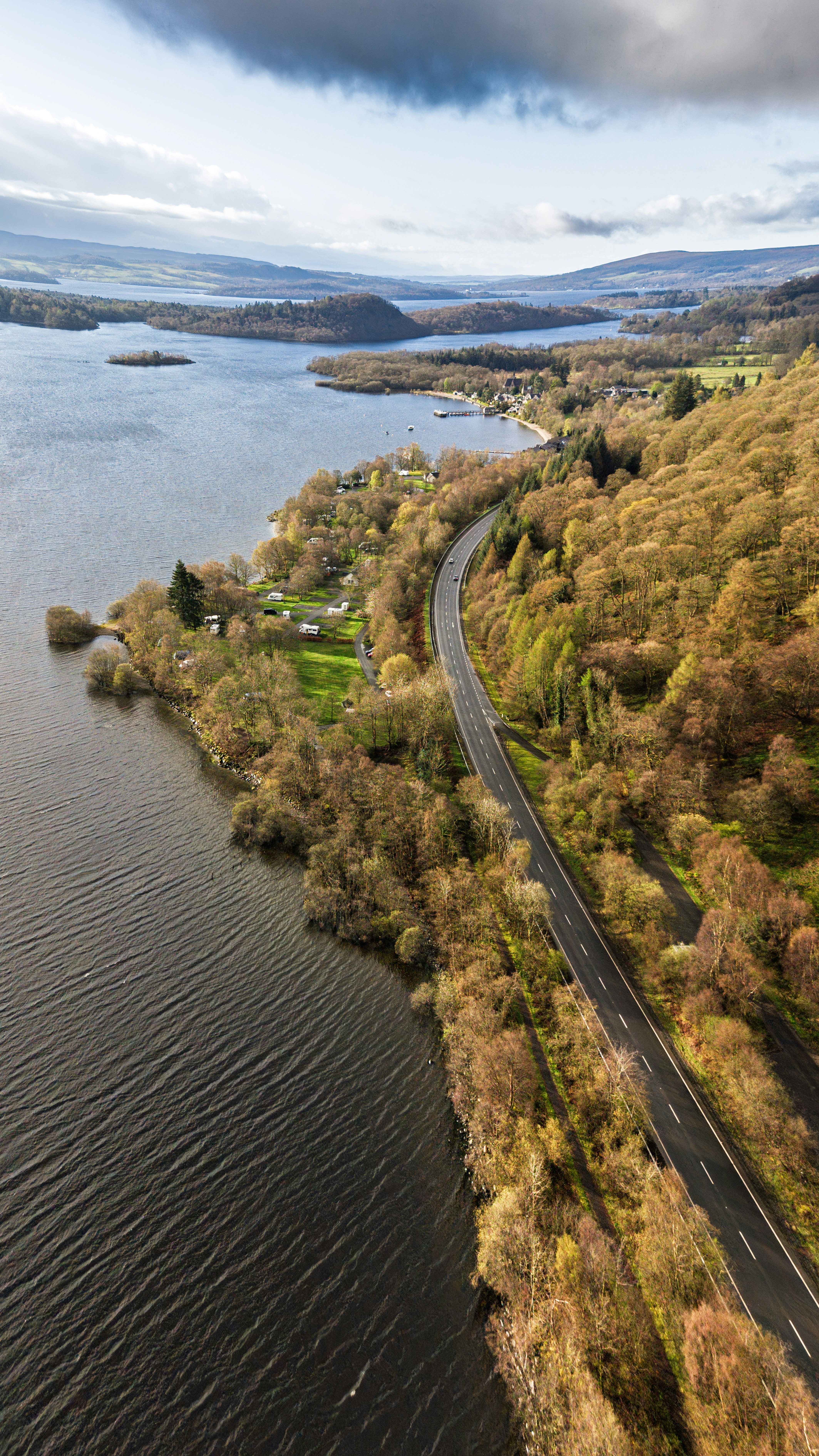 Straße entlang eines Sees mit Herbstbäumen und fernen Hügeln