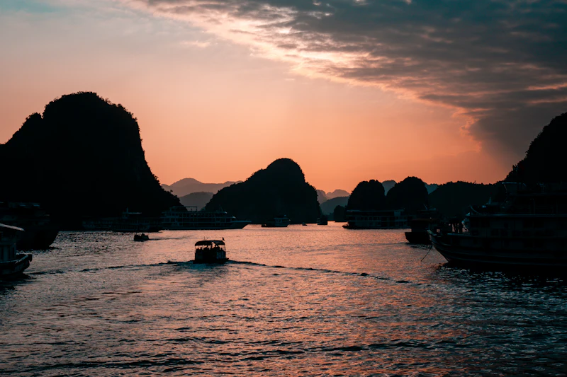 Tour boats crossing Ha Long Bay at sunset between dark limestone karst islands under an orange sky