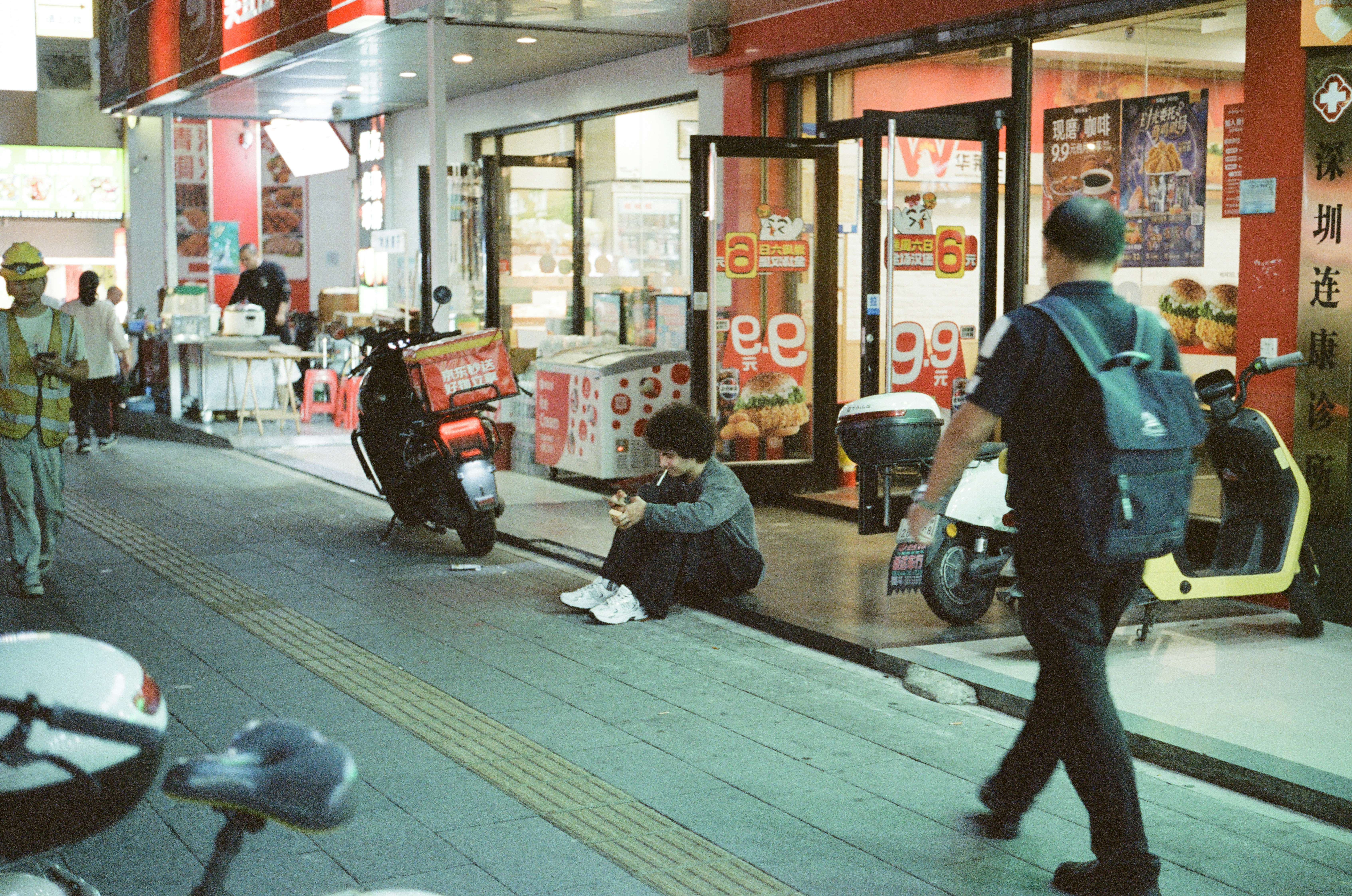 People walking on a city street with shops and scooters.