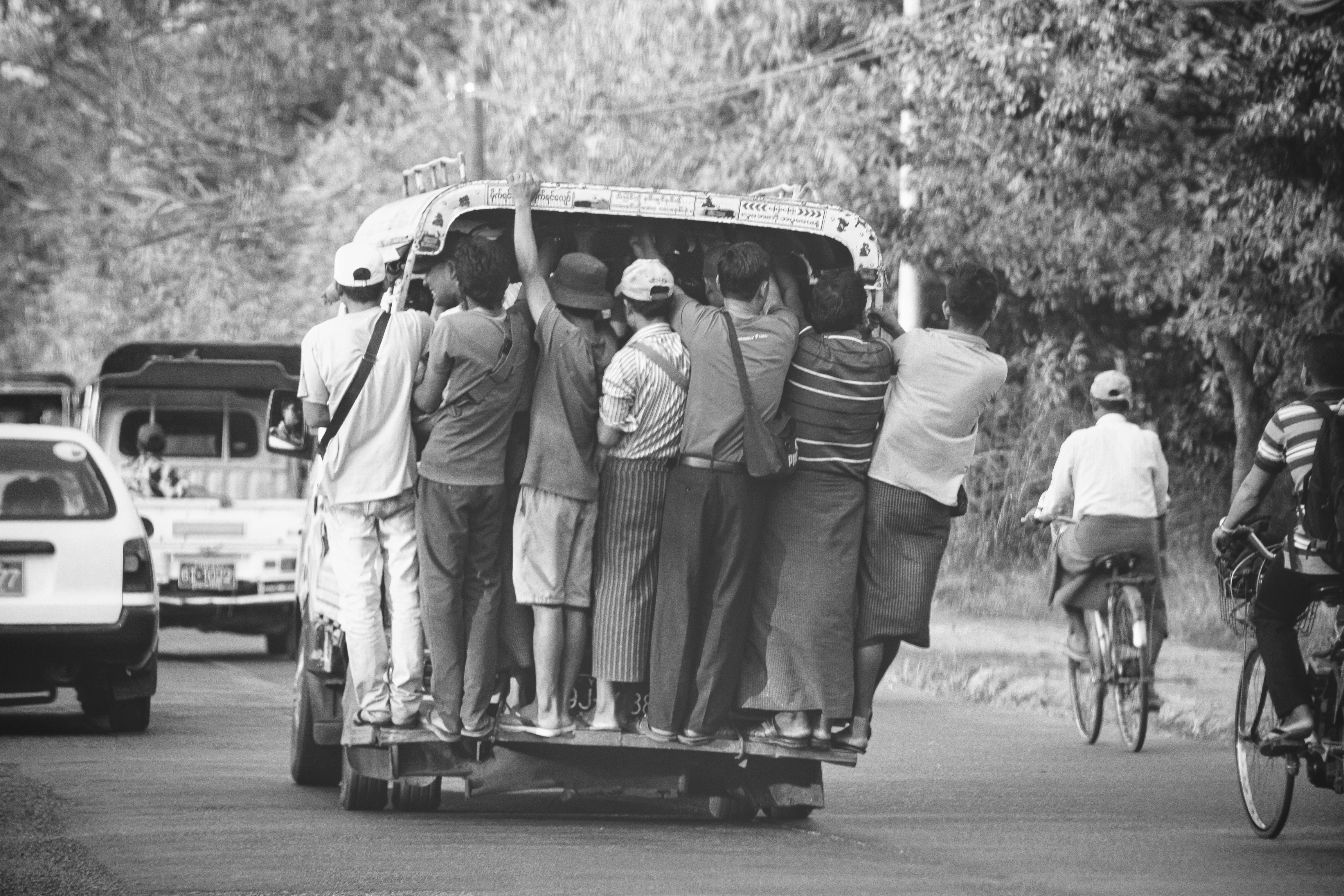 A compelling black and white documentary photograph capturing the daily commute in suburban Yangon, Myanmar. Over a dozen male passengers are packed onto the rear of a small truck-based pickup, holding on for a tight and risky ride. The image depicts the resilience of community life and the challenging transportation reality. The leading lines of passengers contrast against the blurred roadside.