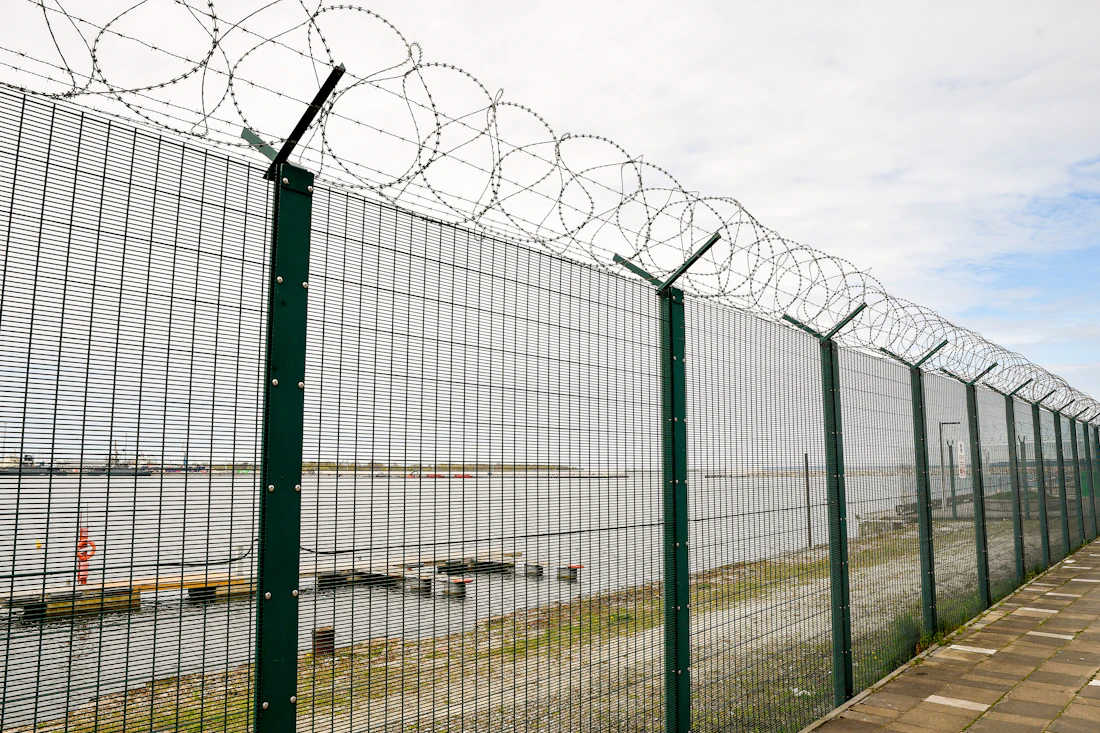 Tall green welded-panel security fence topped with barbed wire