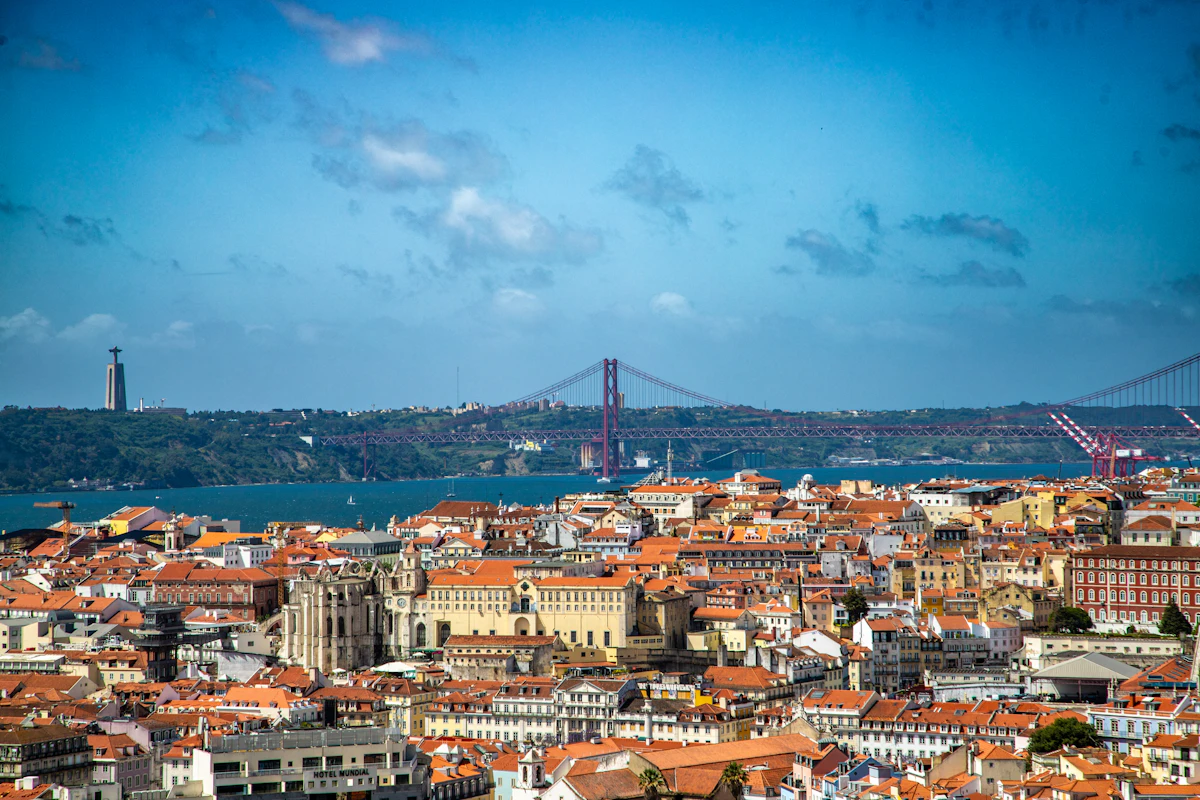 Vista de Lisboa, com edifícios históricos, o rio Tejo e uma ponte ao fundo.