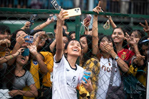 Young woman takes a selfie with cheering crowd behind fence