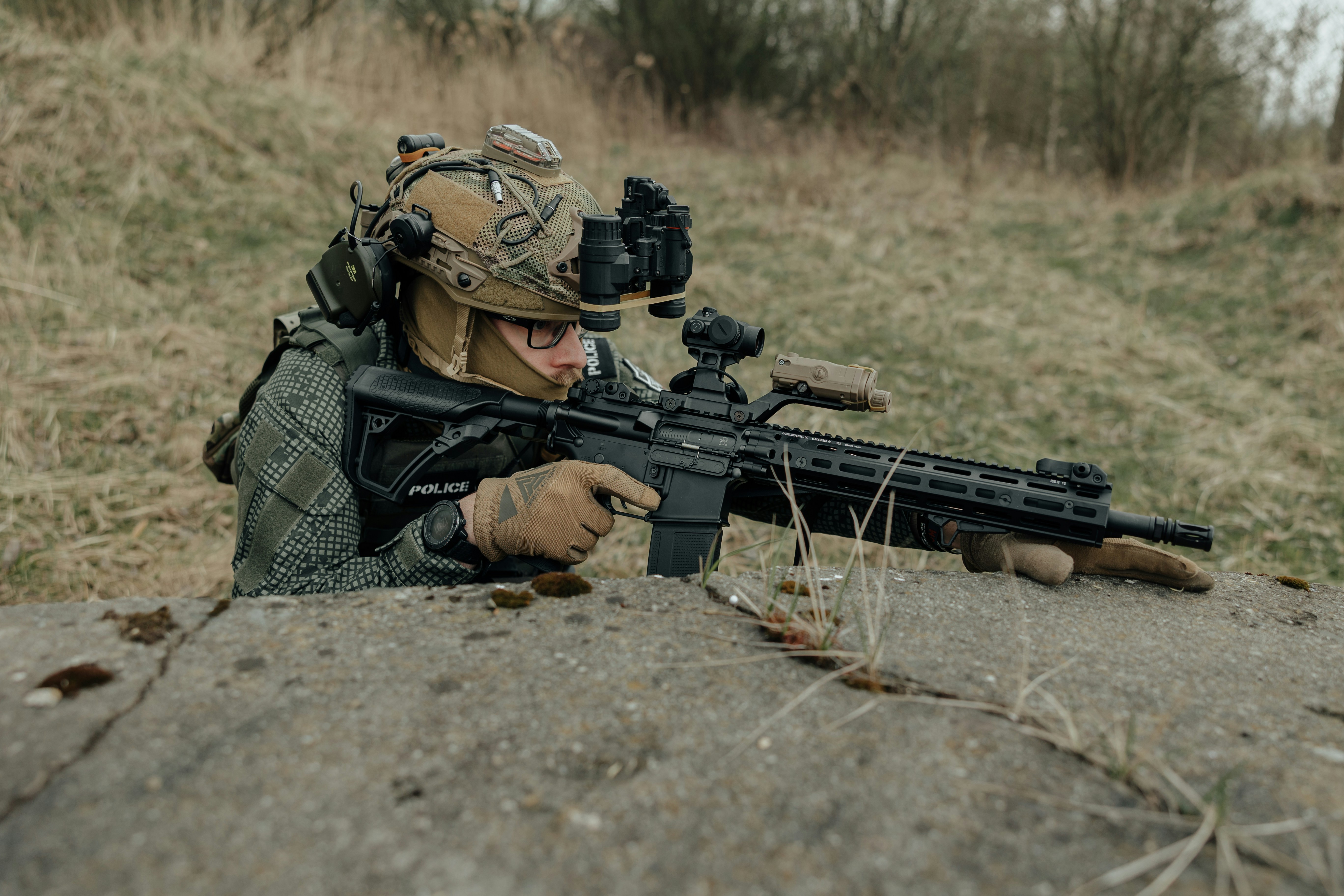 Soldier in tactical gear aiming rifle from behind cover.