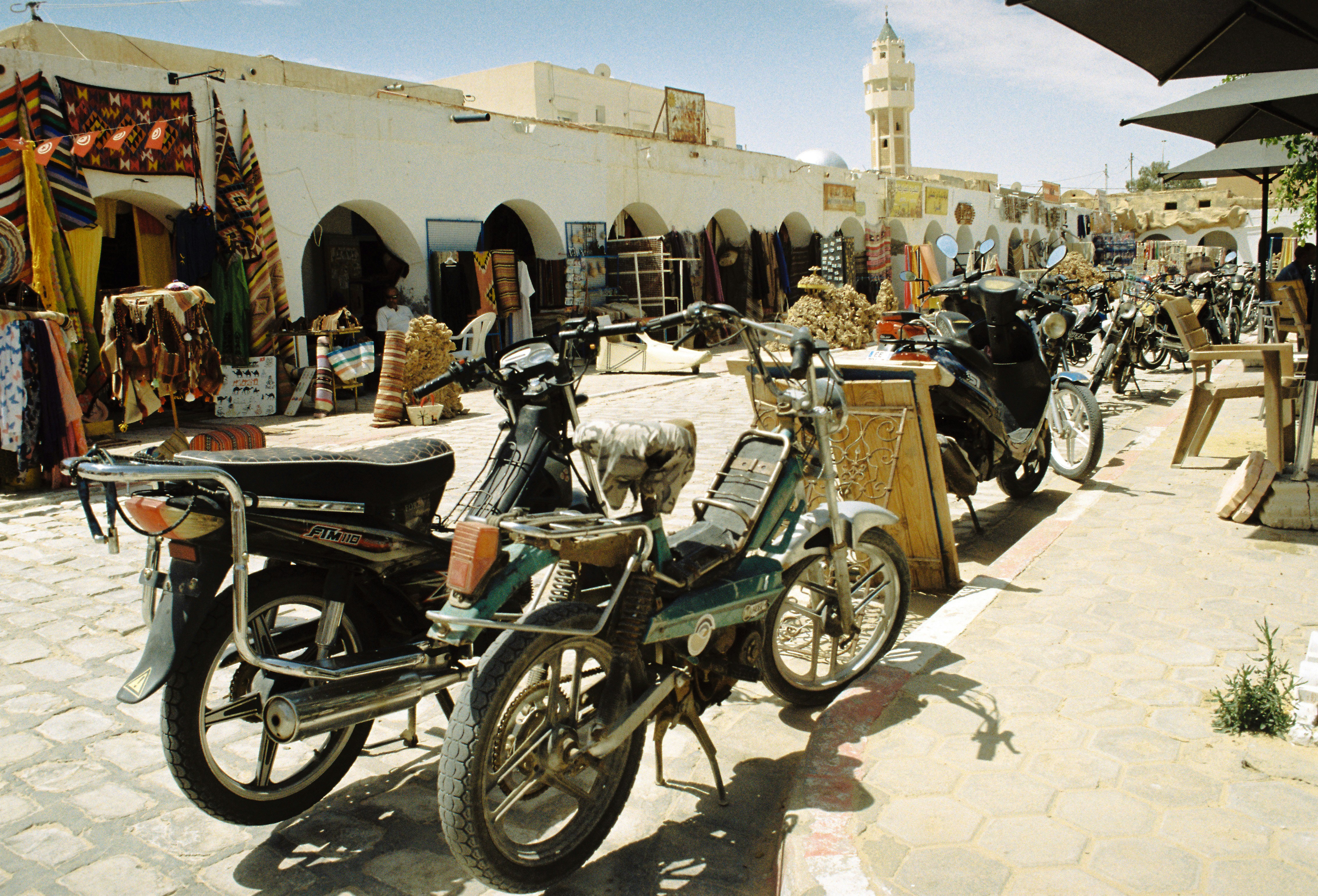 Motorcycles parked on a sunny street with shops.