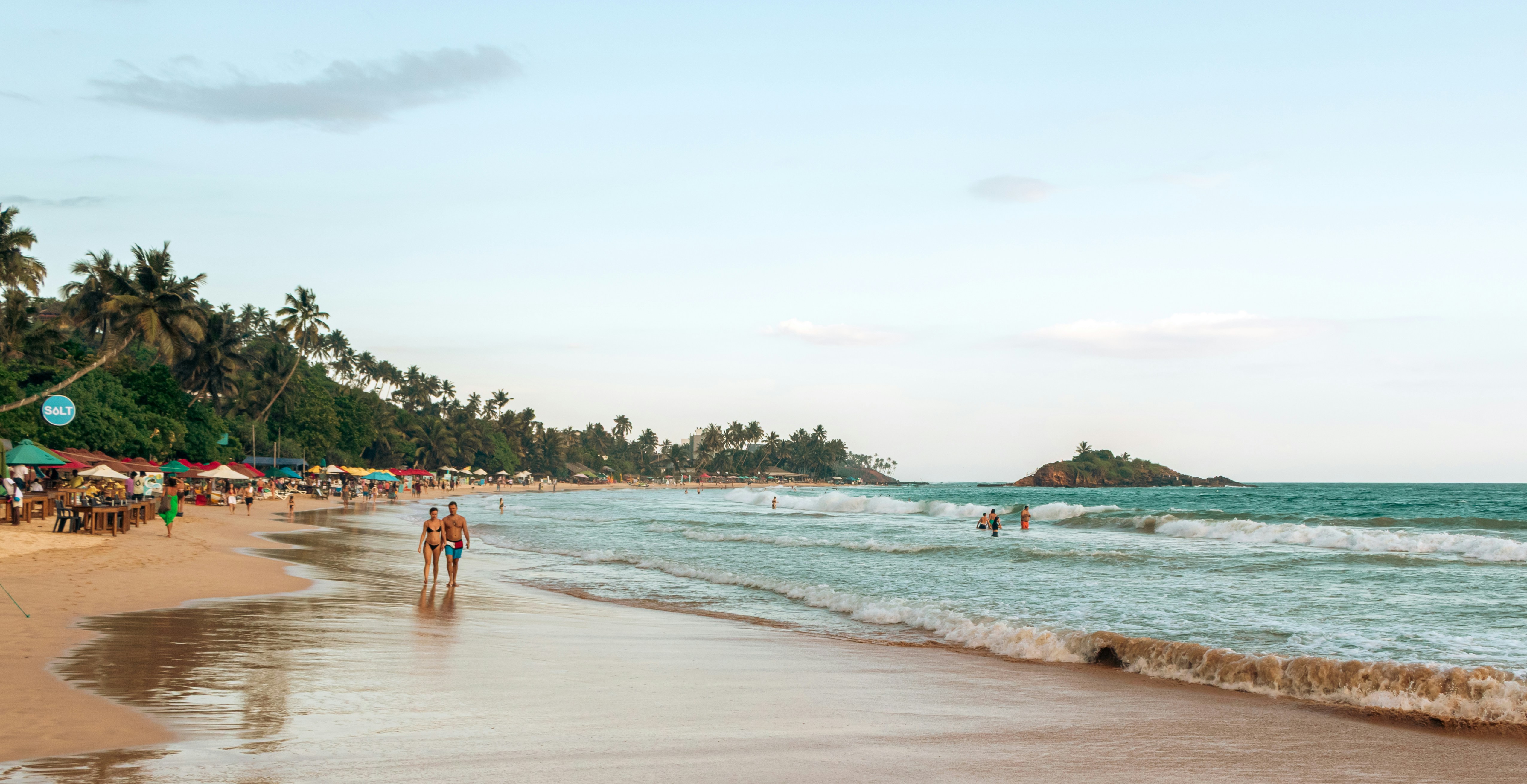People walk along a tropical beach with palm trees.