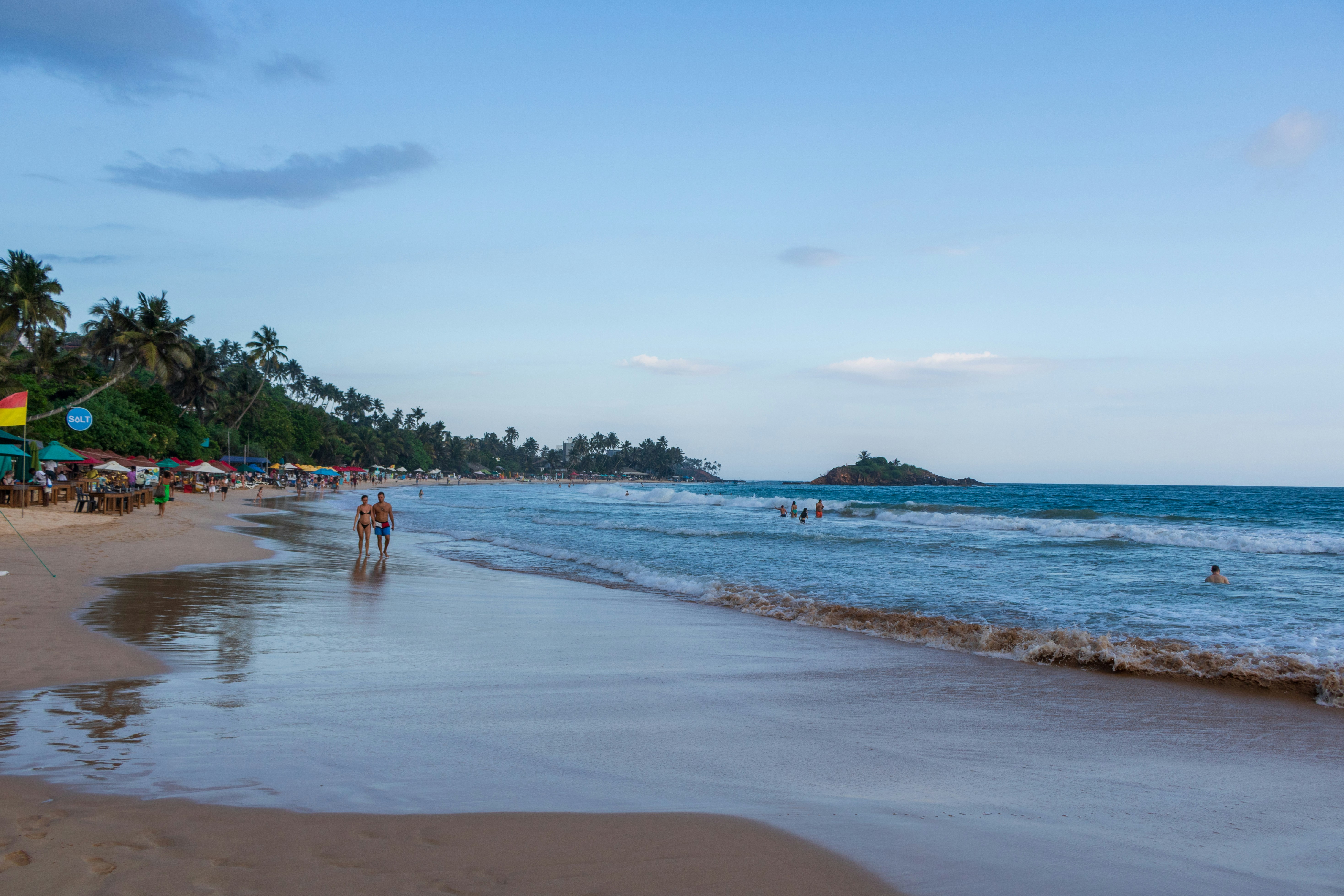 People walk along a tropical beach with palm trees.