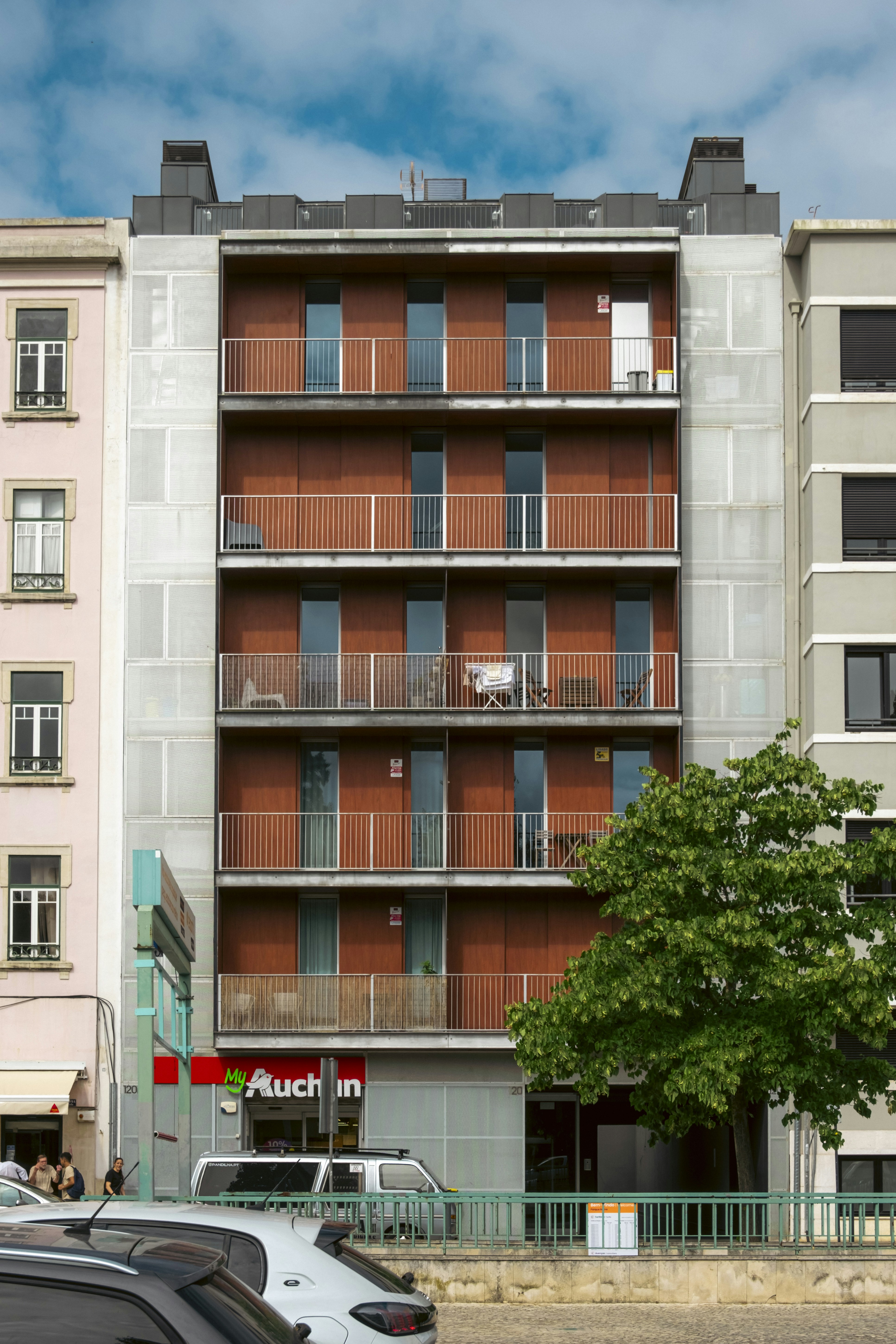 A contemporary residential building in downtown Lisbon with rust-colored metal balconies and geometric symmetry, contrasted against older neighboring architecture and a dramatic blue sky. With Auchan shop on the ground floor. Photo made in Summer 2025. Shot with Fujifilm XM5 and Fujinon XC15-45. // Um edifício residencial contemporâneo no centro de Lisboa com varandas em metal de tom enferrujado e simetria geométrica, contrastando com a arquitetura mais antiga ao lado e um céu azul dramático.