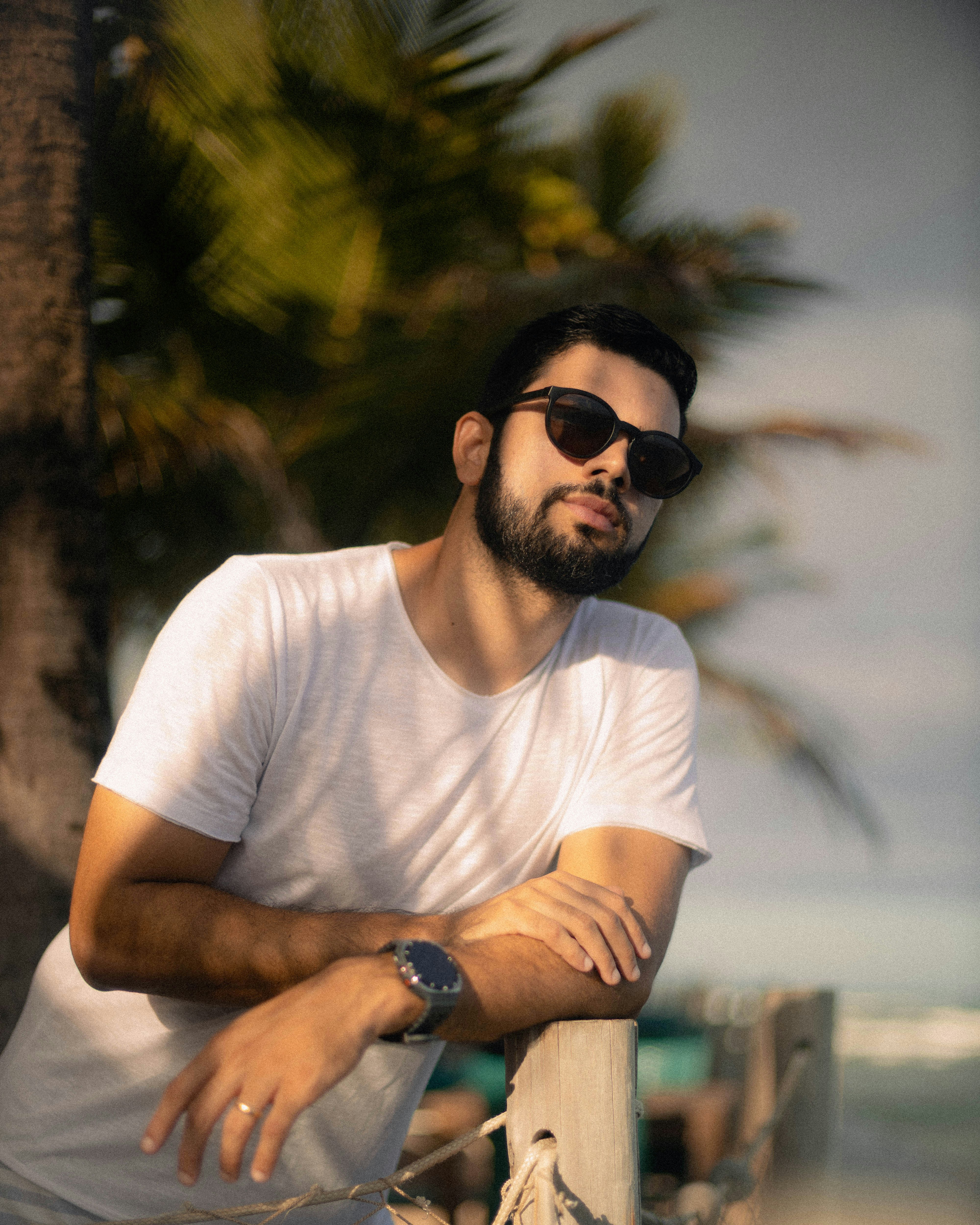 Hombre con gafas de sol apoyado en un poste de madera en la playa.