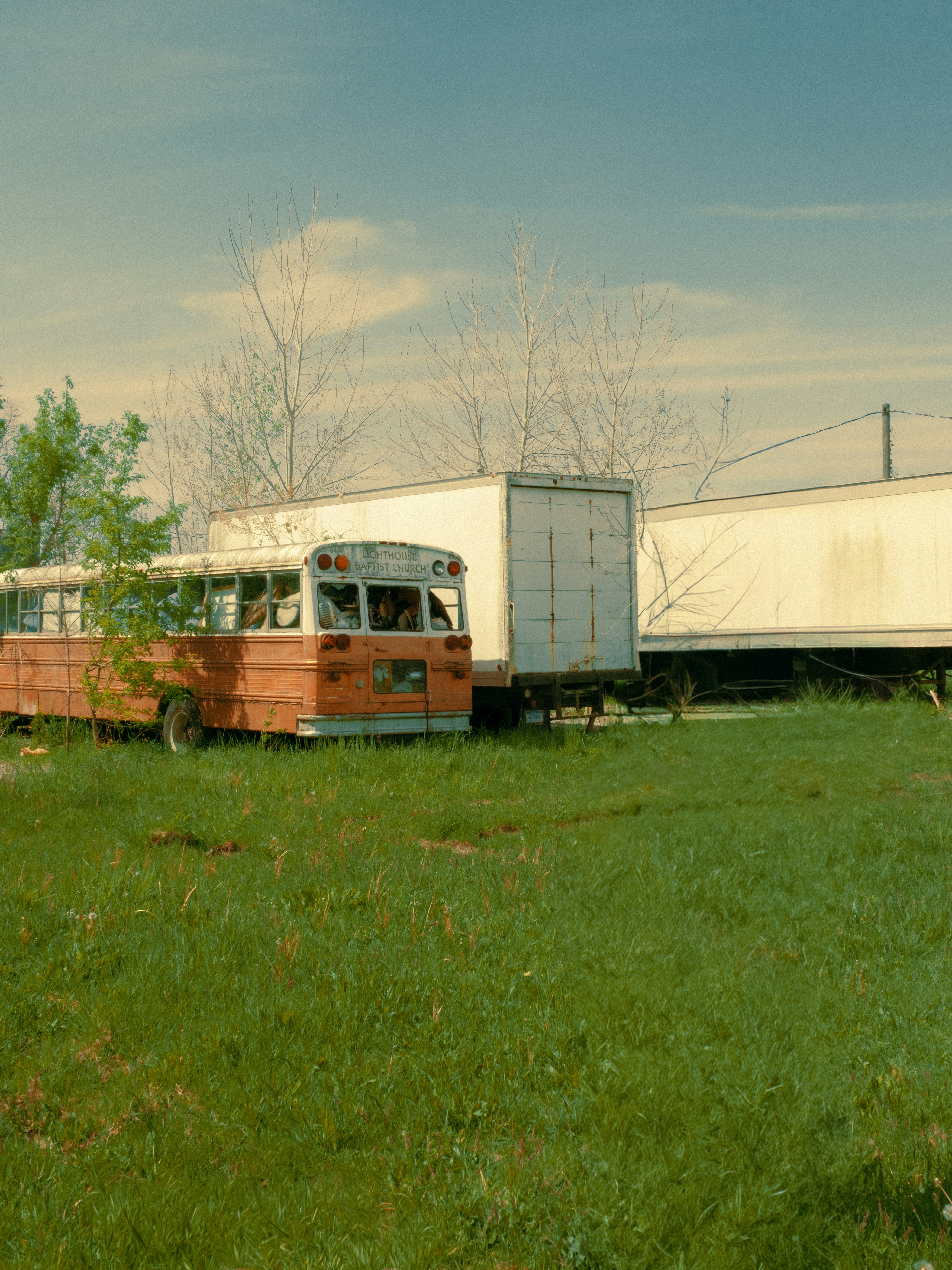 Rusty orange school bus in a field with white semi-trailers.