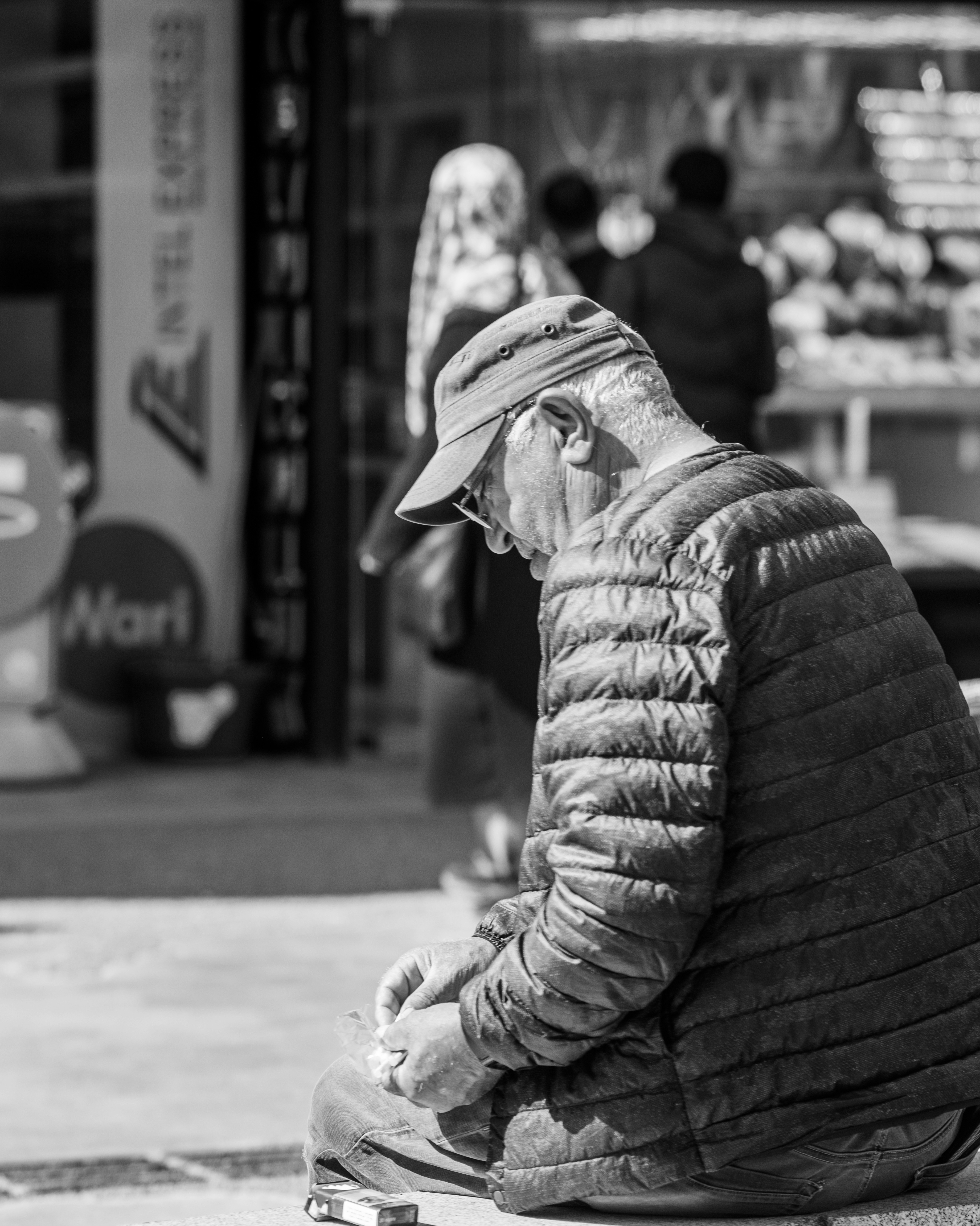 パッファージャケットとキャップをかぶった高齢の男性が外に座っている。