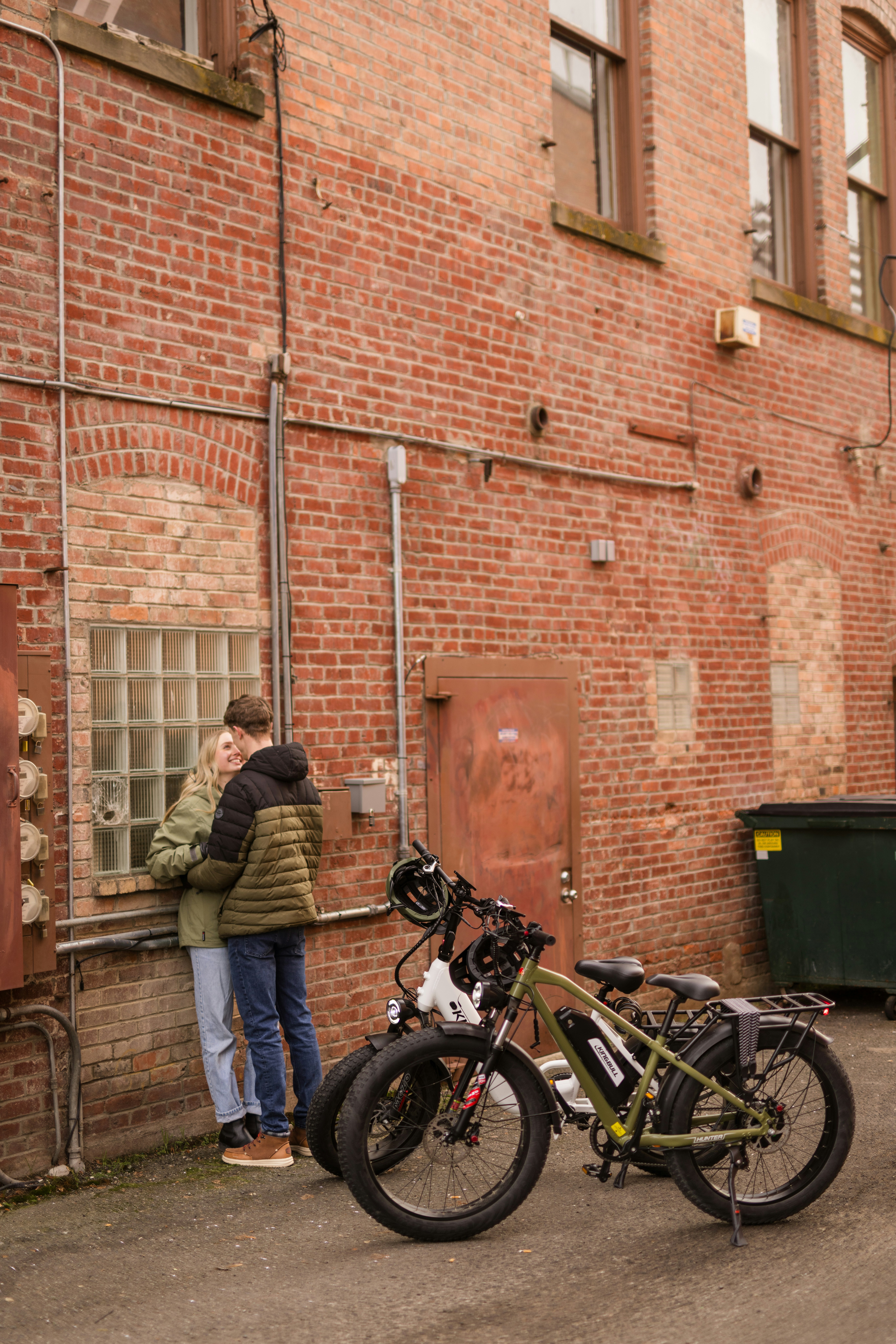 Casal abraçado perto de uma parede de tijolos com bicicletas elétricas.