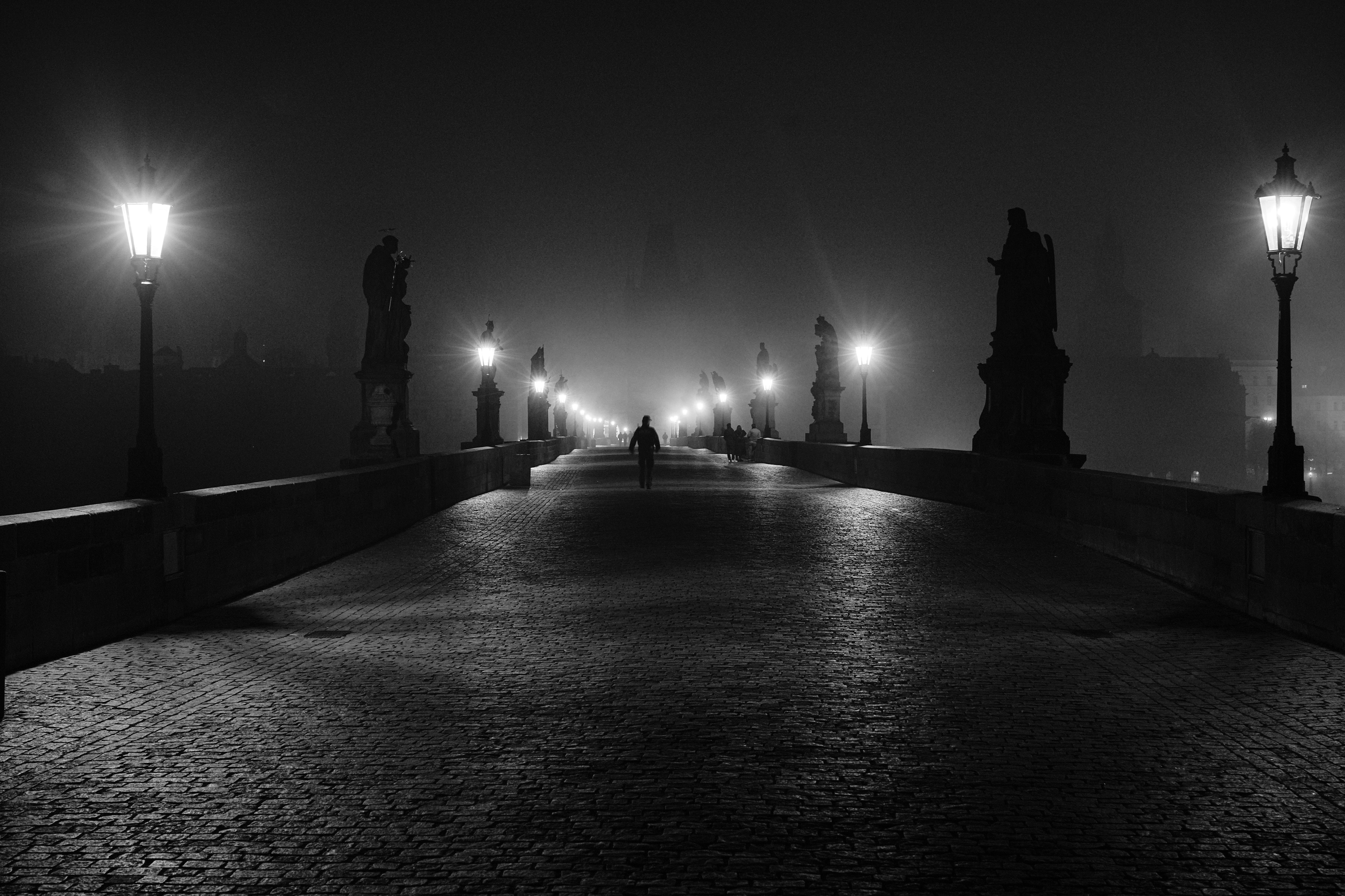 A lone person walks on a foggy bridge at night.