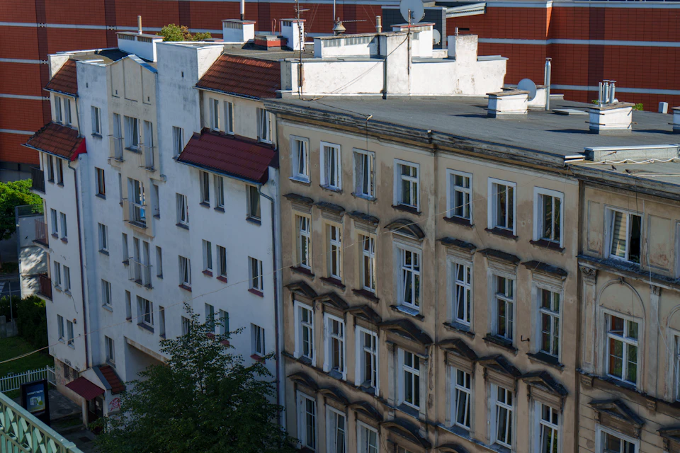 The image shows a cityscape with two-story buildings, one white and one brown, situated on a street corner. The white building has a red roof and is topped by a chimney, while the brown building features a flat roof and also has a chimney. Both buildings have multiple windows that reflect the sunlight. In front of the brown building, there's a tree with lush green leaves.
