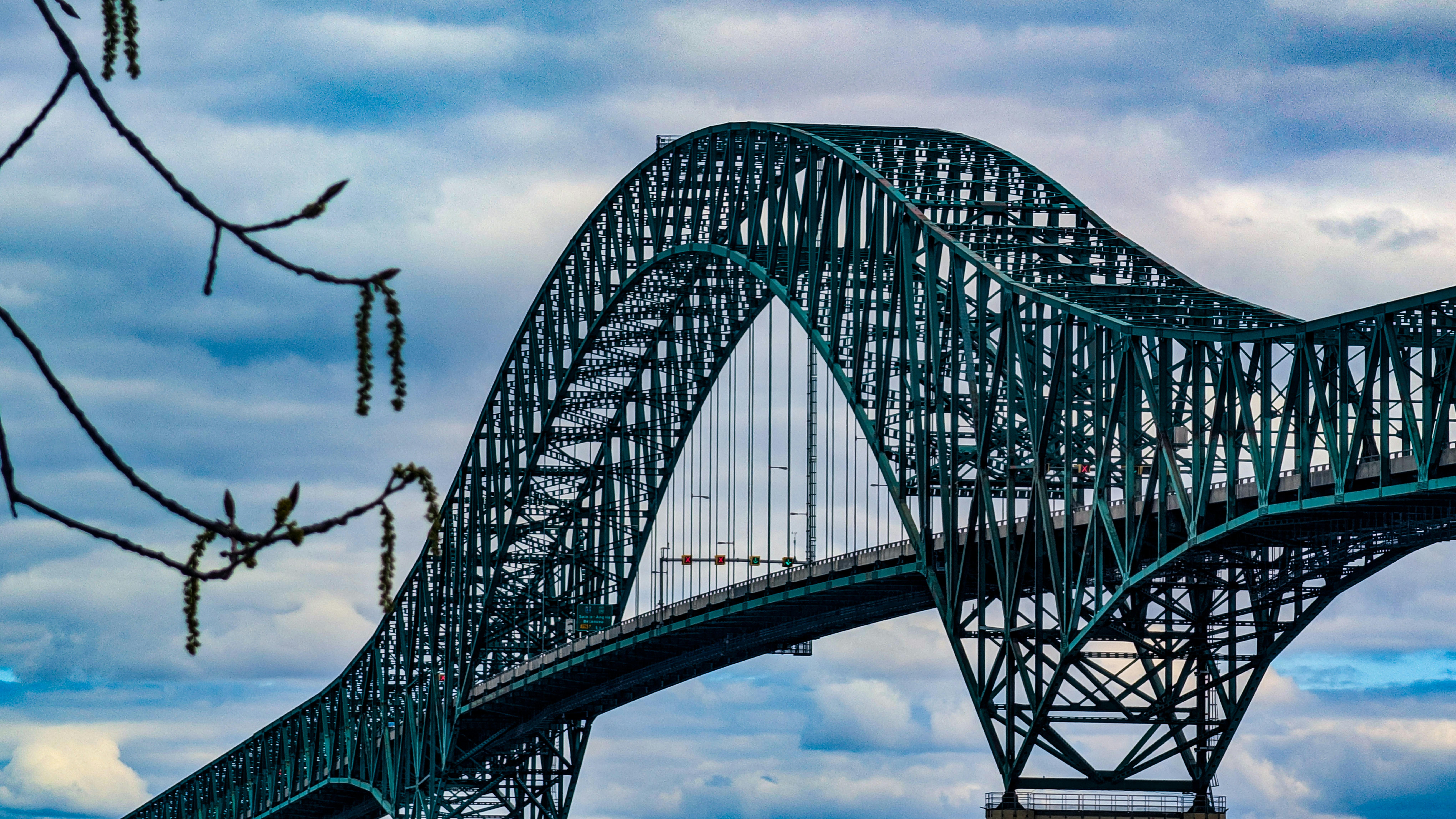 Teal steel arch bridge dominates the frame with dramatic clouds and tree branches in the foreground.