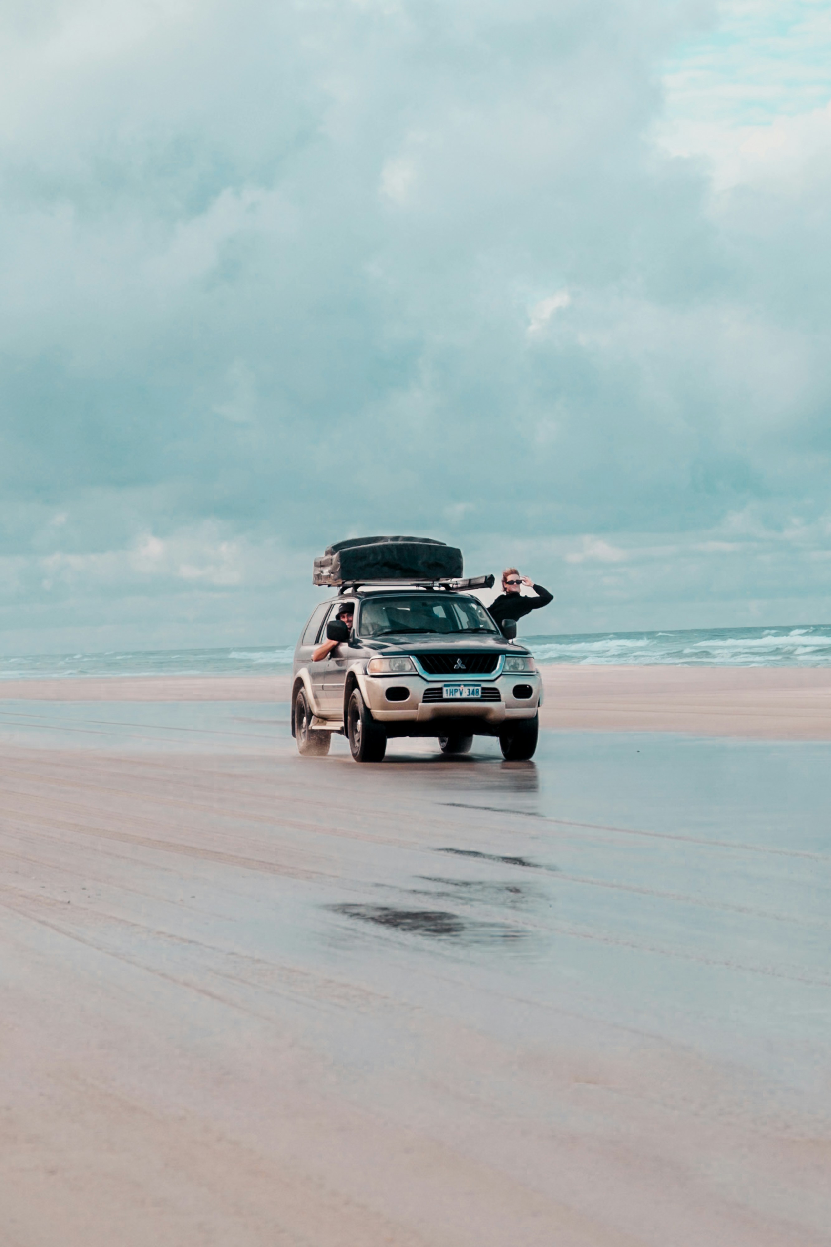 A Mitsubishi Pajero drives along a wide beach with wet sand reflecting the sky under dramatic Australian clouds.
