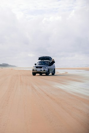 Silver and dark blue SUV driving across a wide, wet sand beach under a cloudy sky, a black soft-sided load strapped to its roof rack