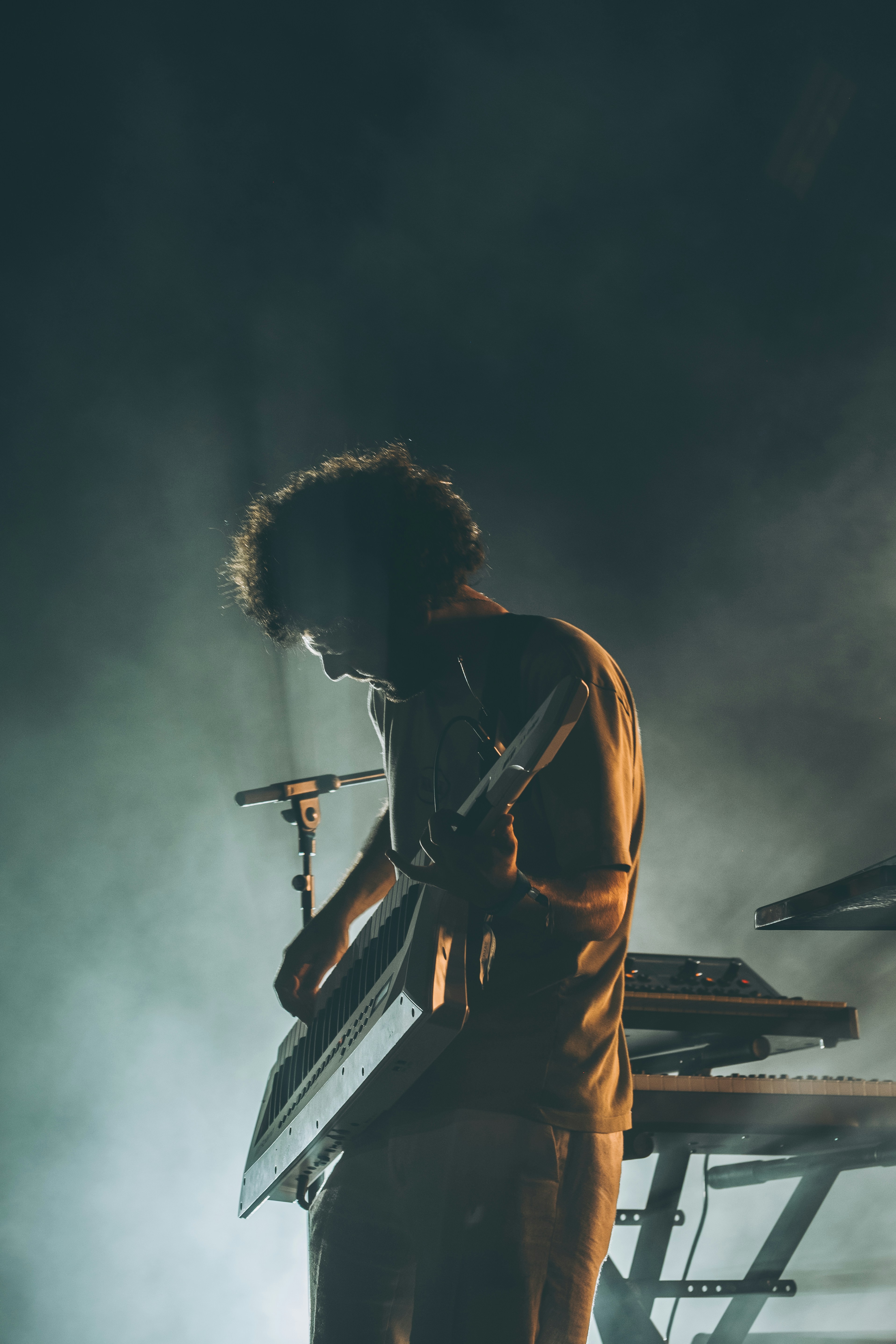 Curly-haired musician plays keytar on a smoky stage.