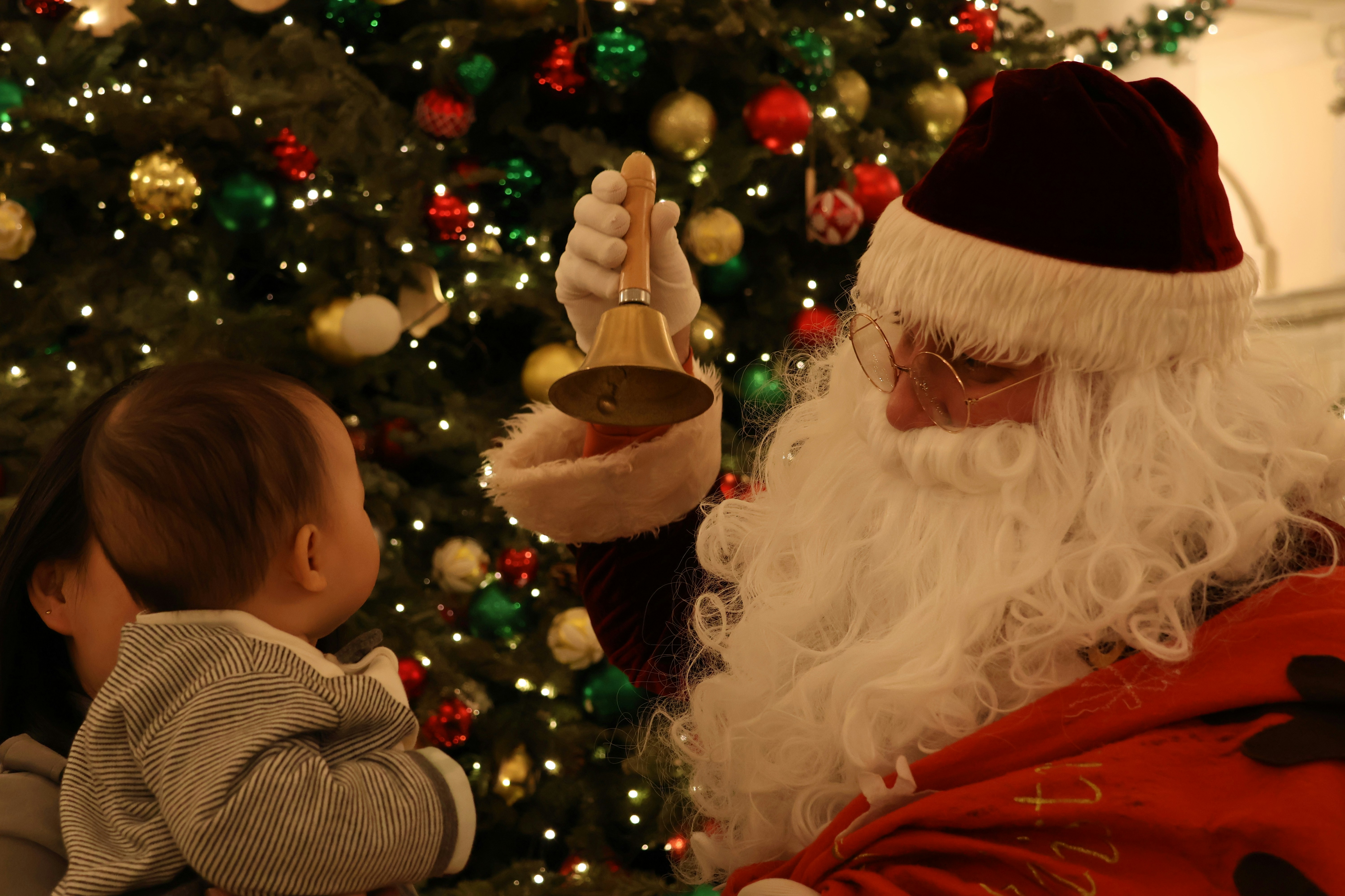 O Papai Noel toca um sino para um bebê perto da árvore de Natal.