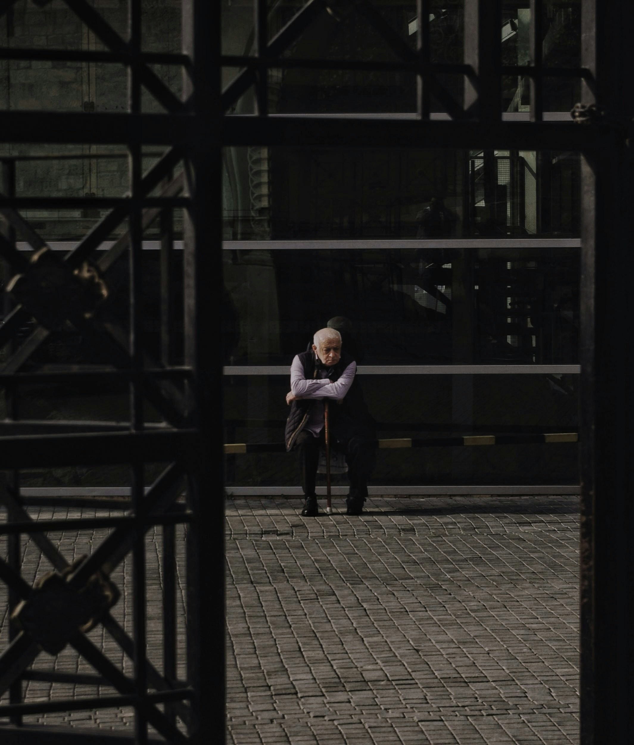 An elderly person sits alone behind a dark metal gate.