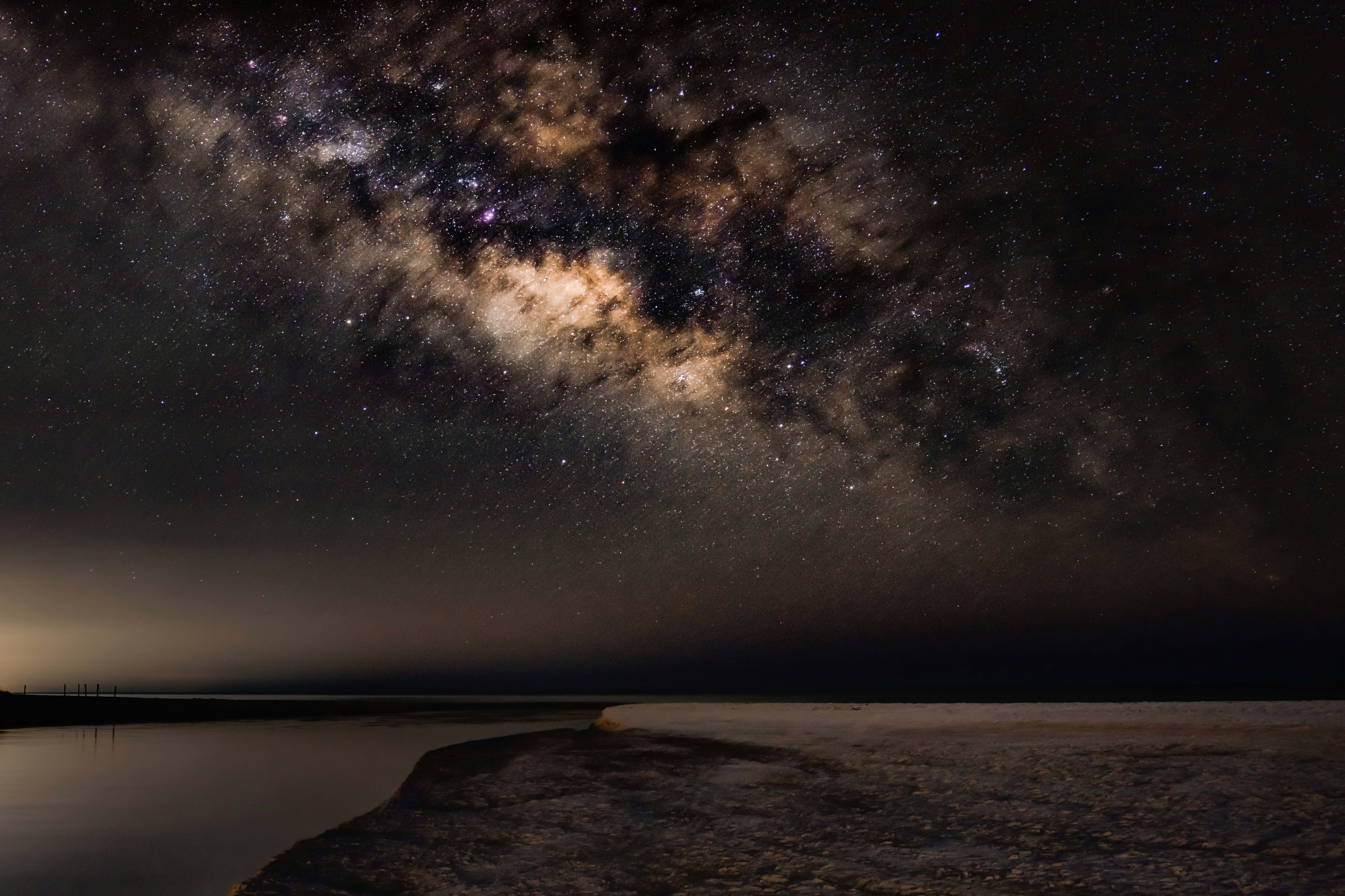 The milky way galaxy arches over a dark, calm landscape.