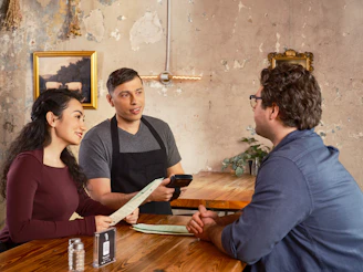 A waiter interacts with two customers at a restaurant table.