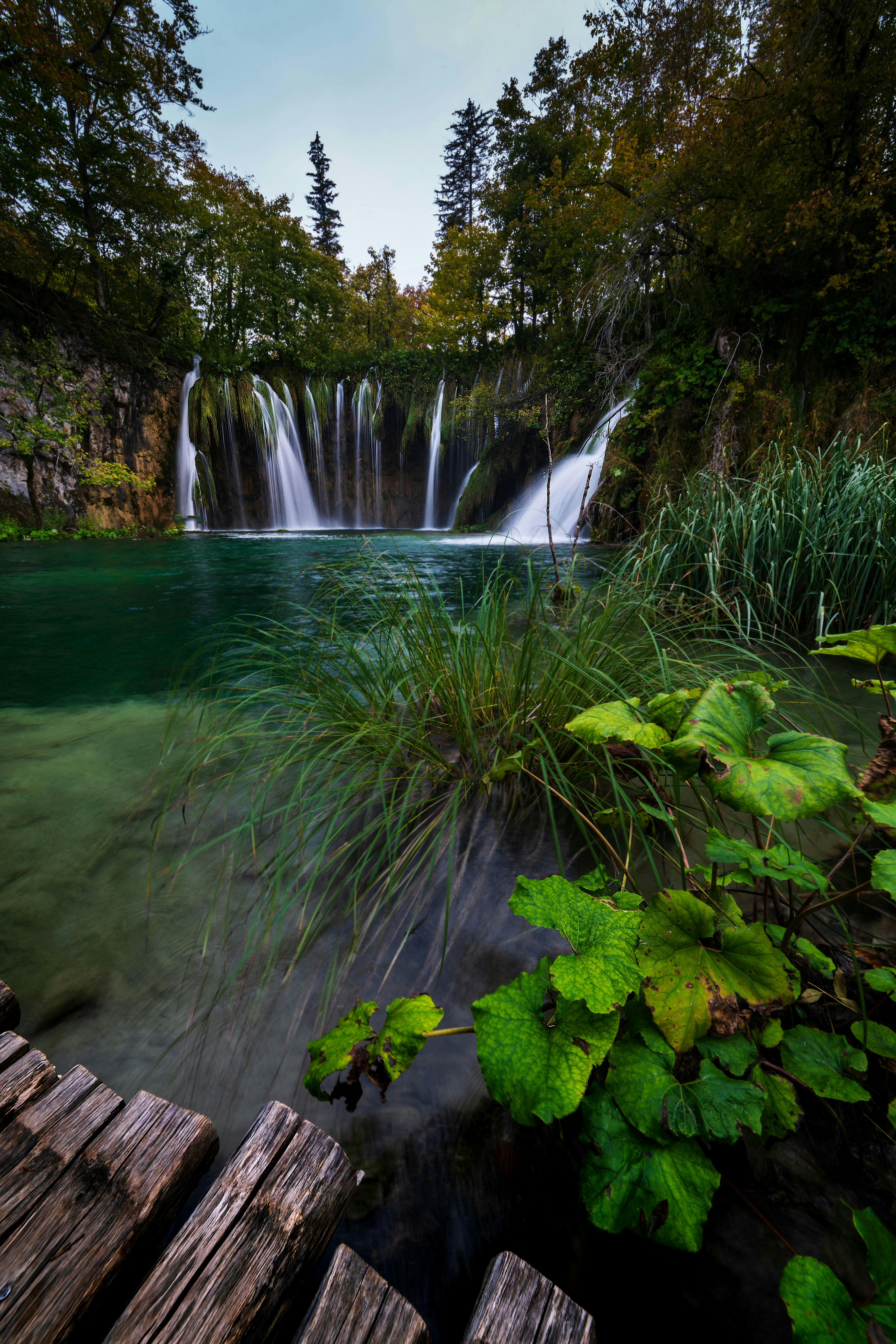 Cascading waterfalls flow into green water beside a wooden path.