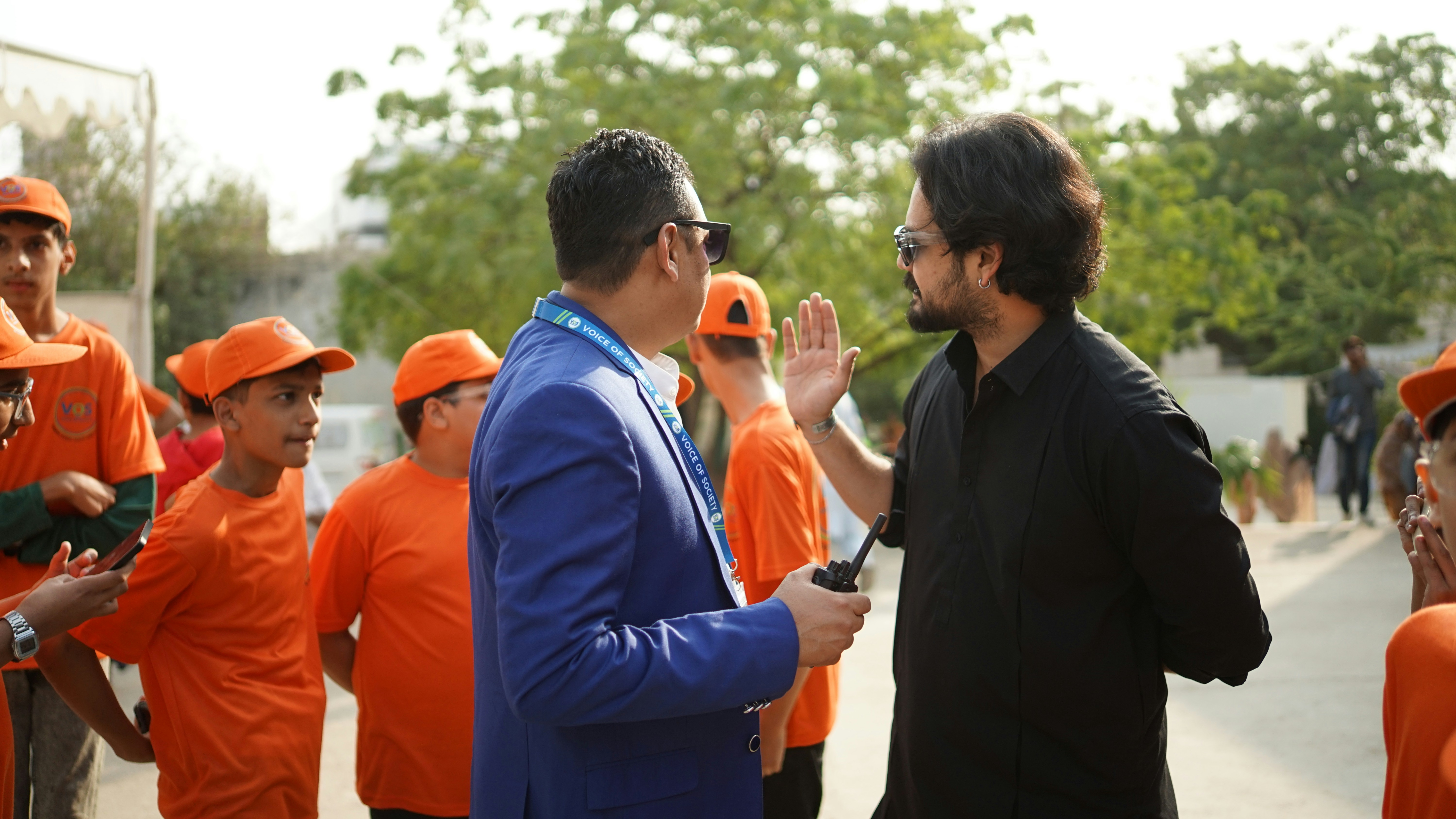 Two men talking with children in orange shirts behind them