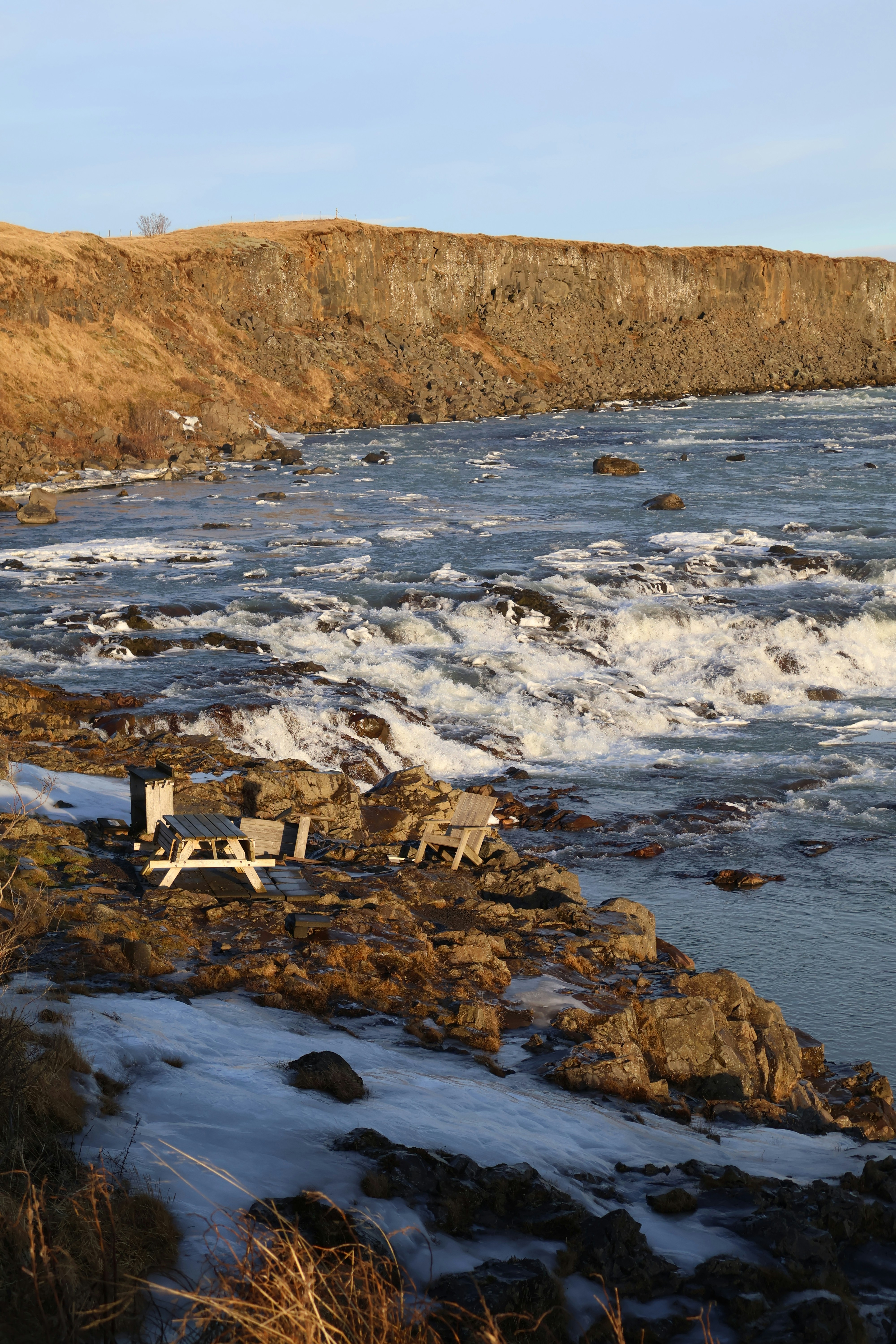A river with rapids, snowy bank, picnic table, and cliff.