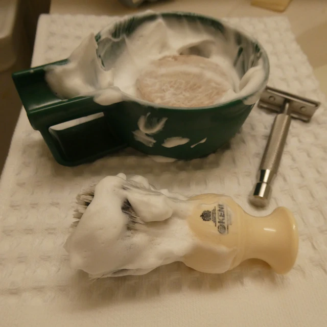 A shaving bowl filled with white lather next to a shaving brush and a razor