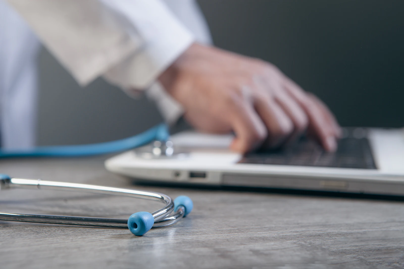 A doctor typing on a laptop with a stethoscope resting on the desk beside it