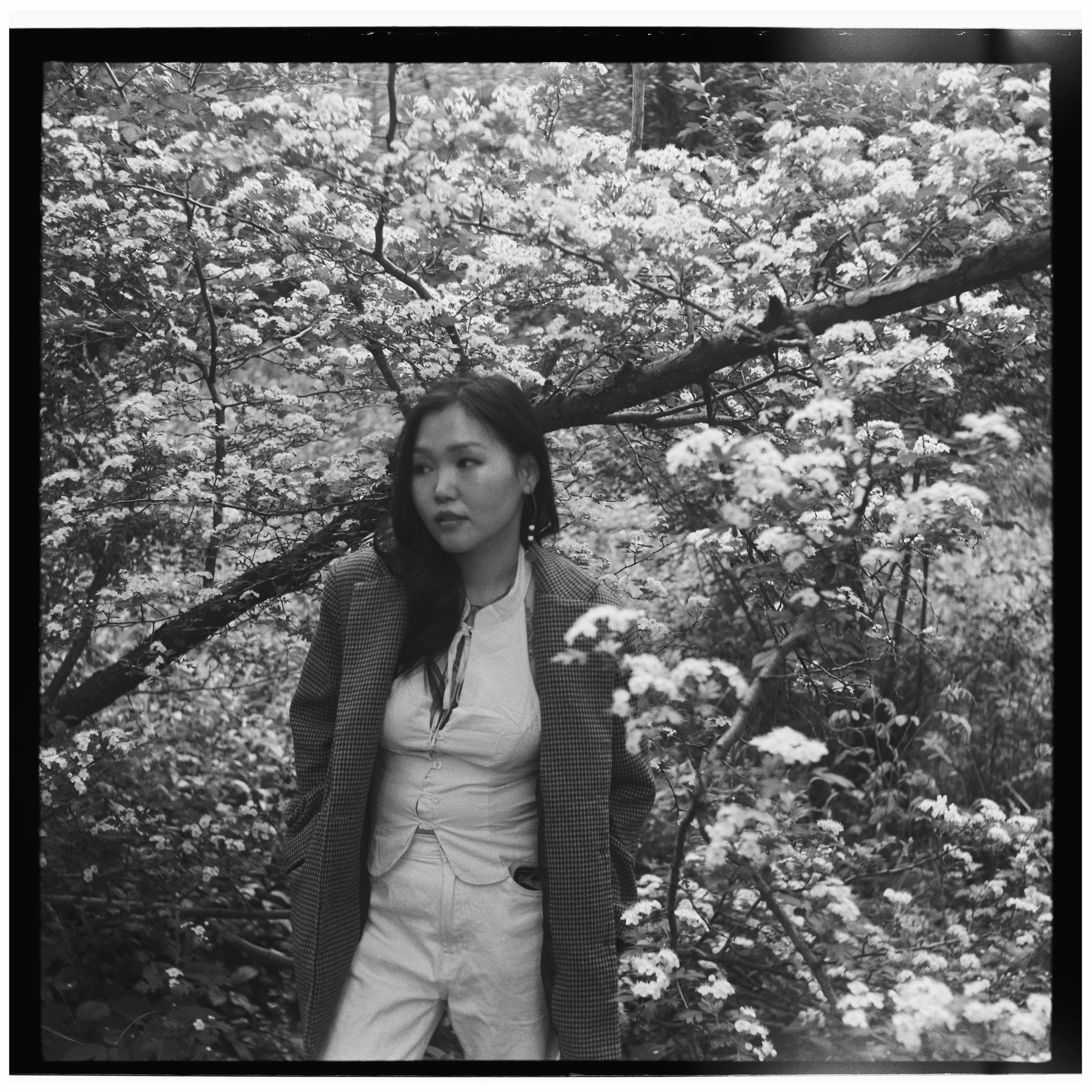Woman in a coat stands amidst blooming trees.