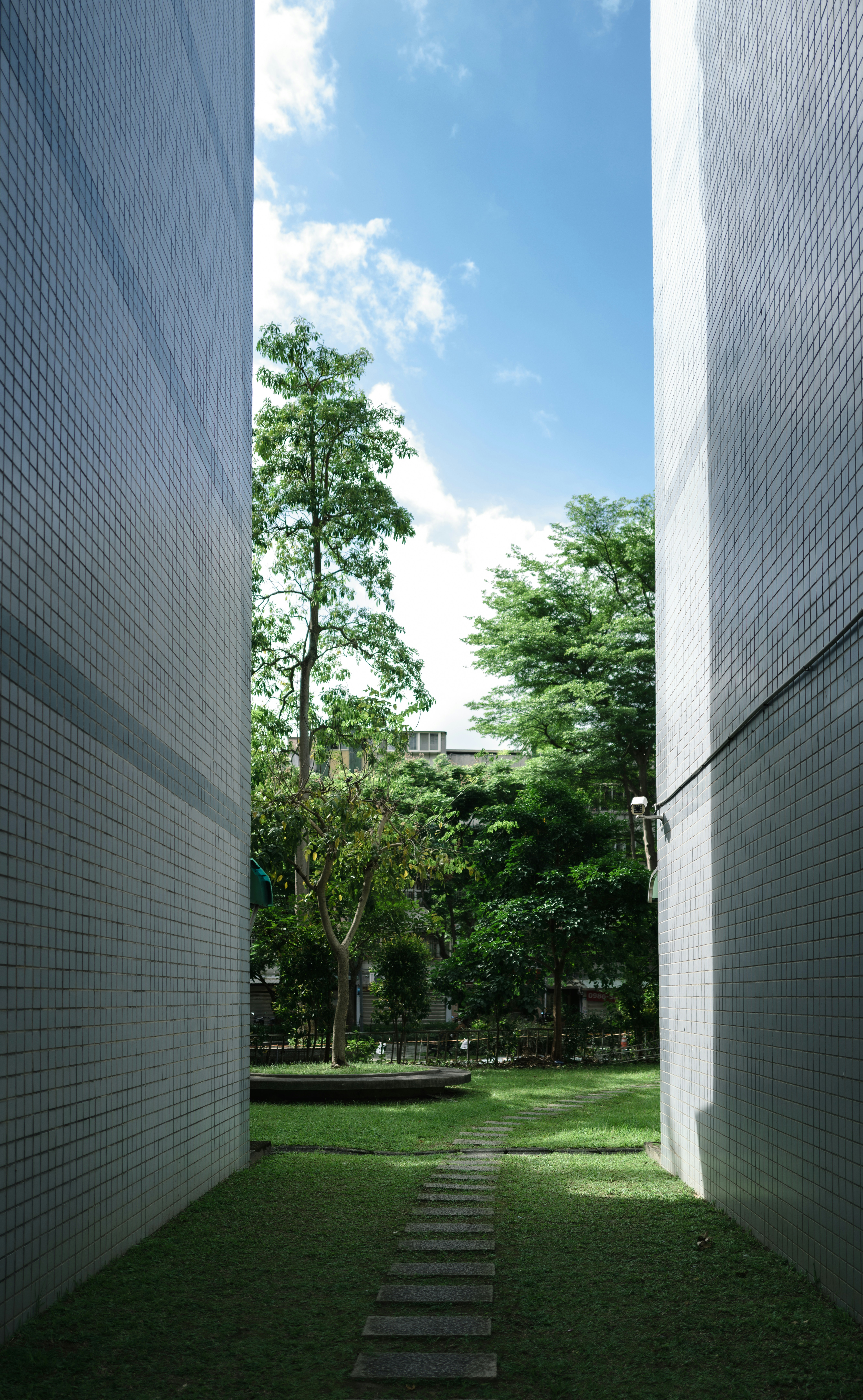 A path leads through green grass between buildings to trees.