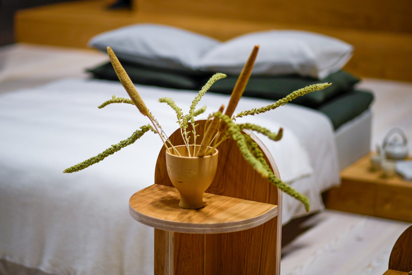 Japandi bedroom detail with dried foxtail grasses in a hand-carved face vase on a curved oak side table next to a low wood platform bed