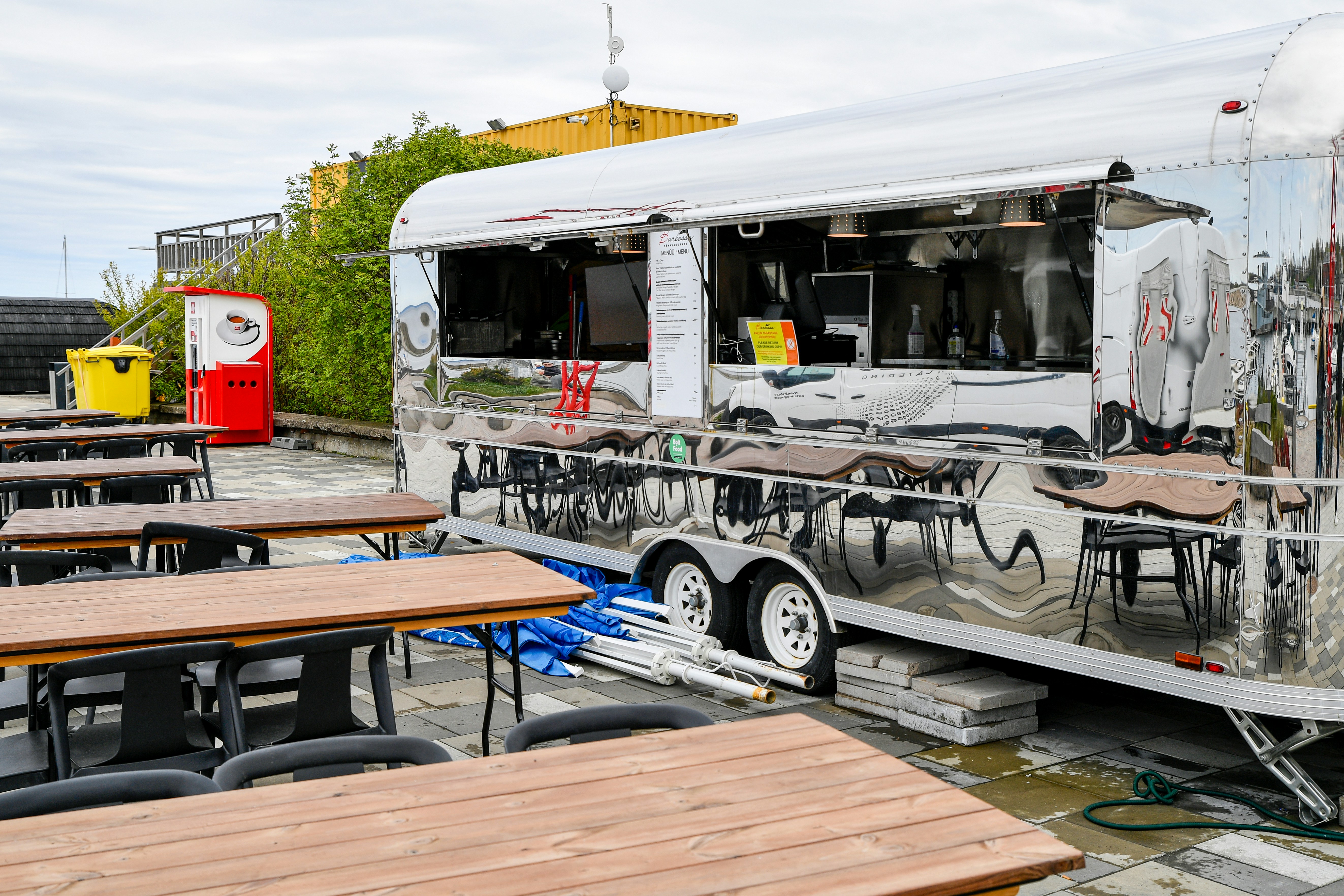 Chrome retro food truck trailer with a mirror finish parked at an outdoor cafe terrace with wooden tables under an overcast sky.