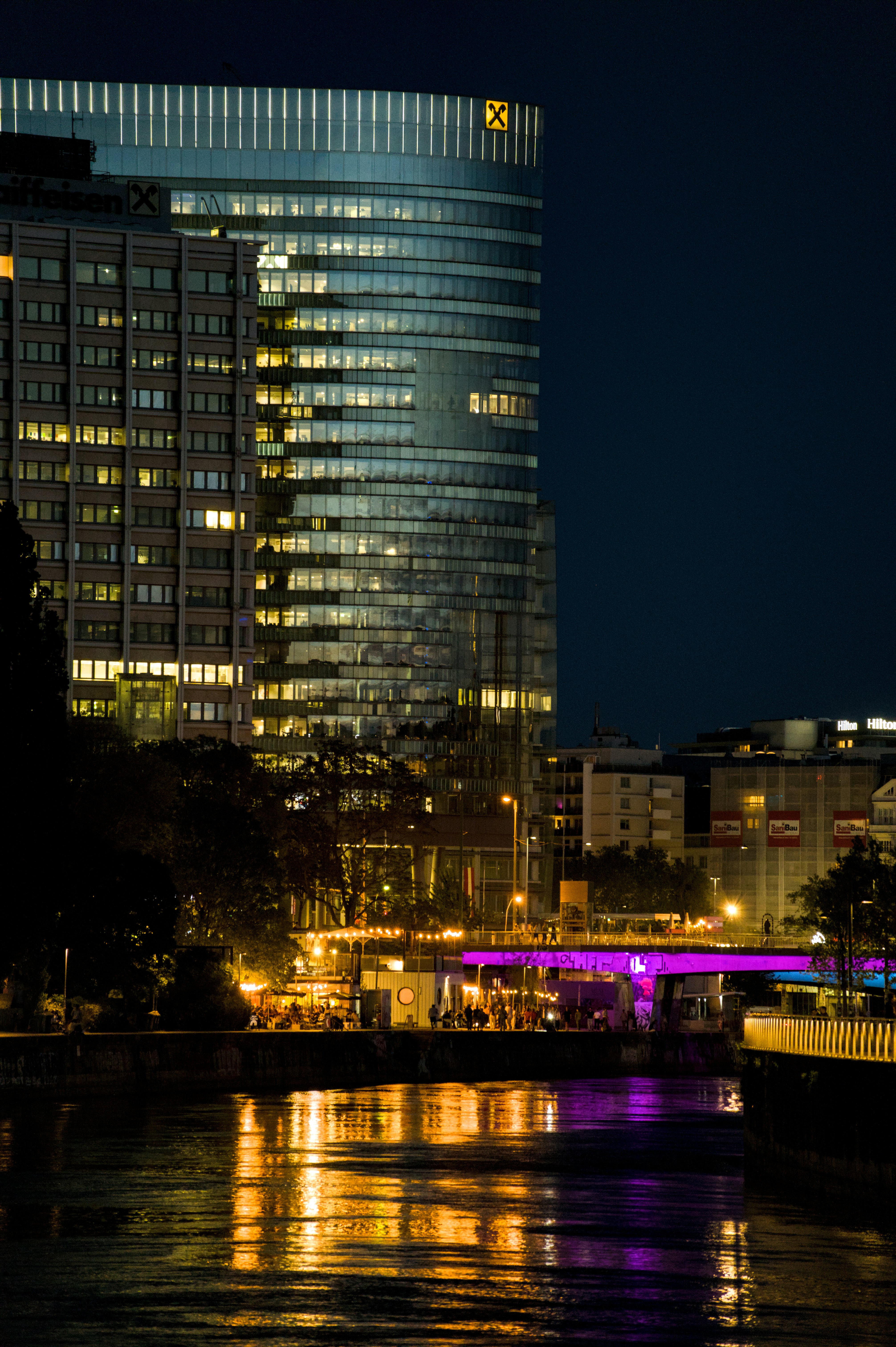 Illuminated modern buildings, river, and purple bridge at night.