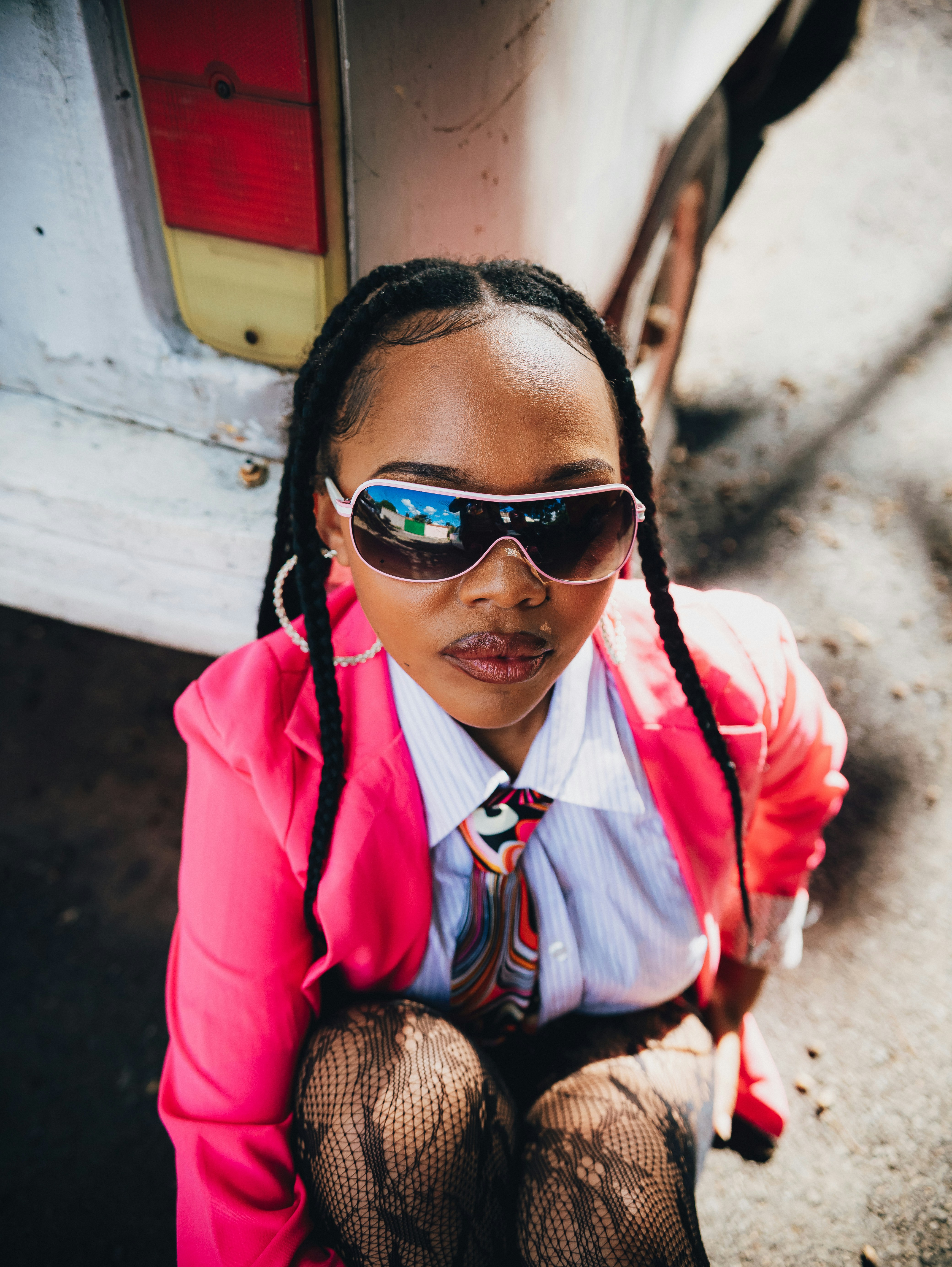 Uma jovem de blazer rosa e óculos escuros sentada ao ar livre.