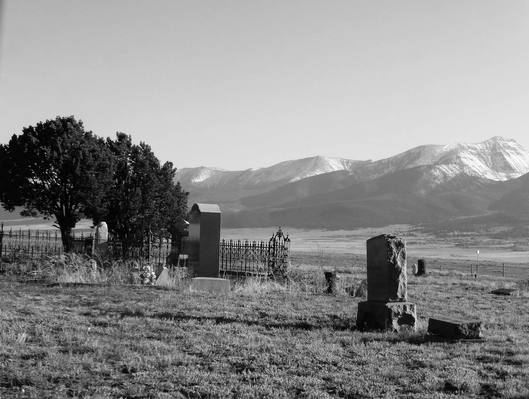 A small rural cemetery of old stones and an iron fence on open grassland with snow-covered mountains behind, in black and white