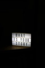 A glowing light box sign asks "what is your story".