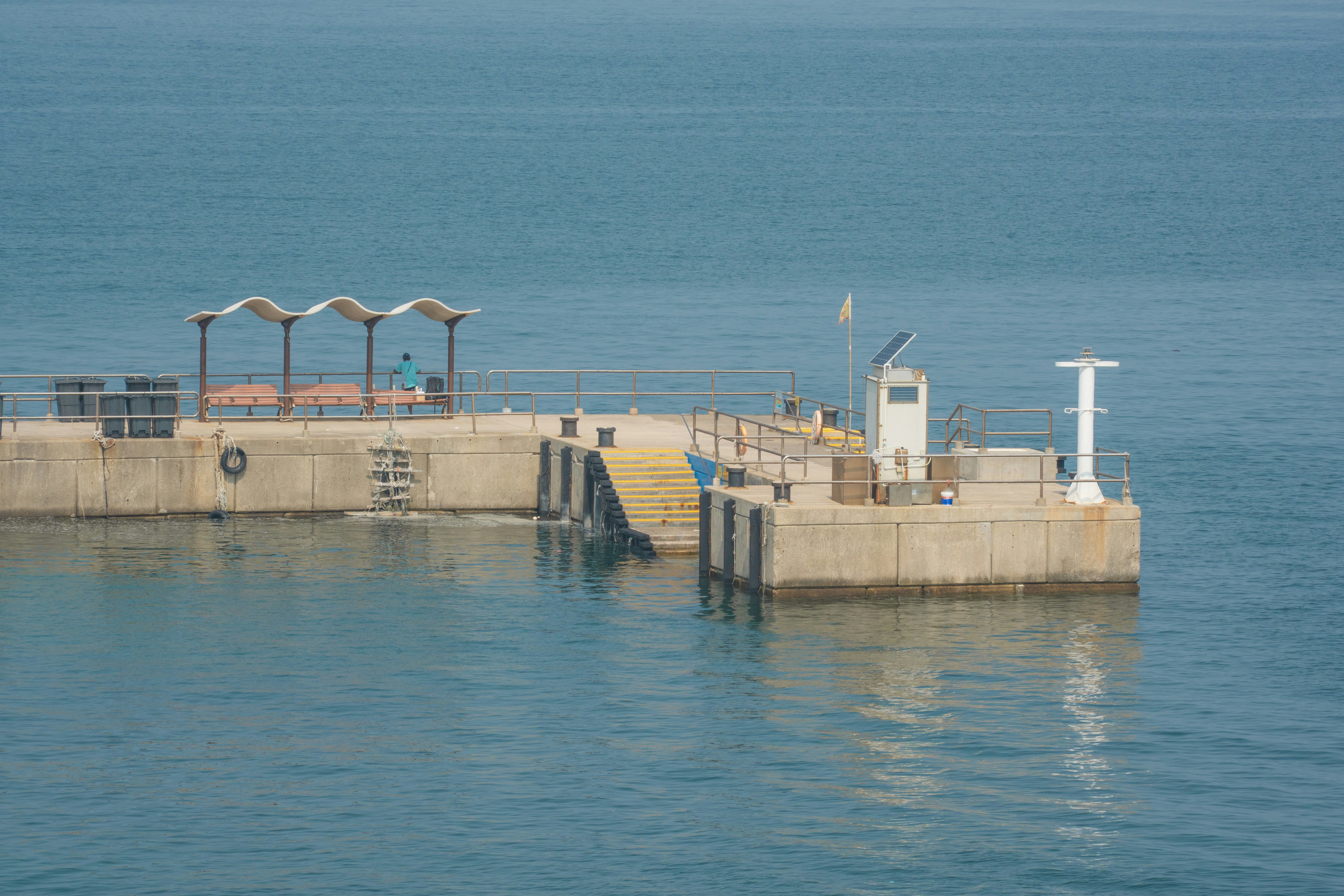 Concrete pier with building and white lighthouse in blue water.