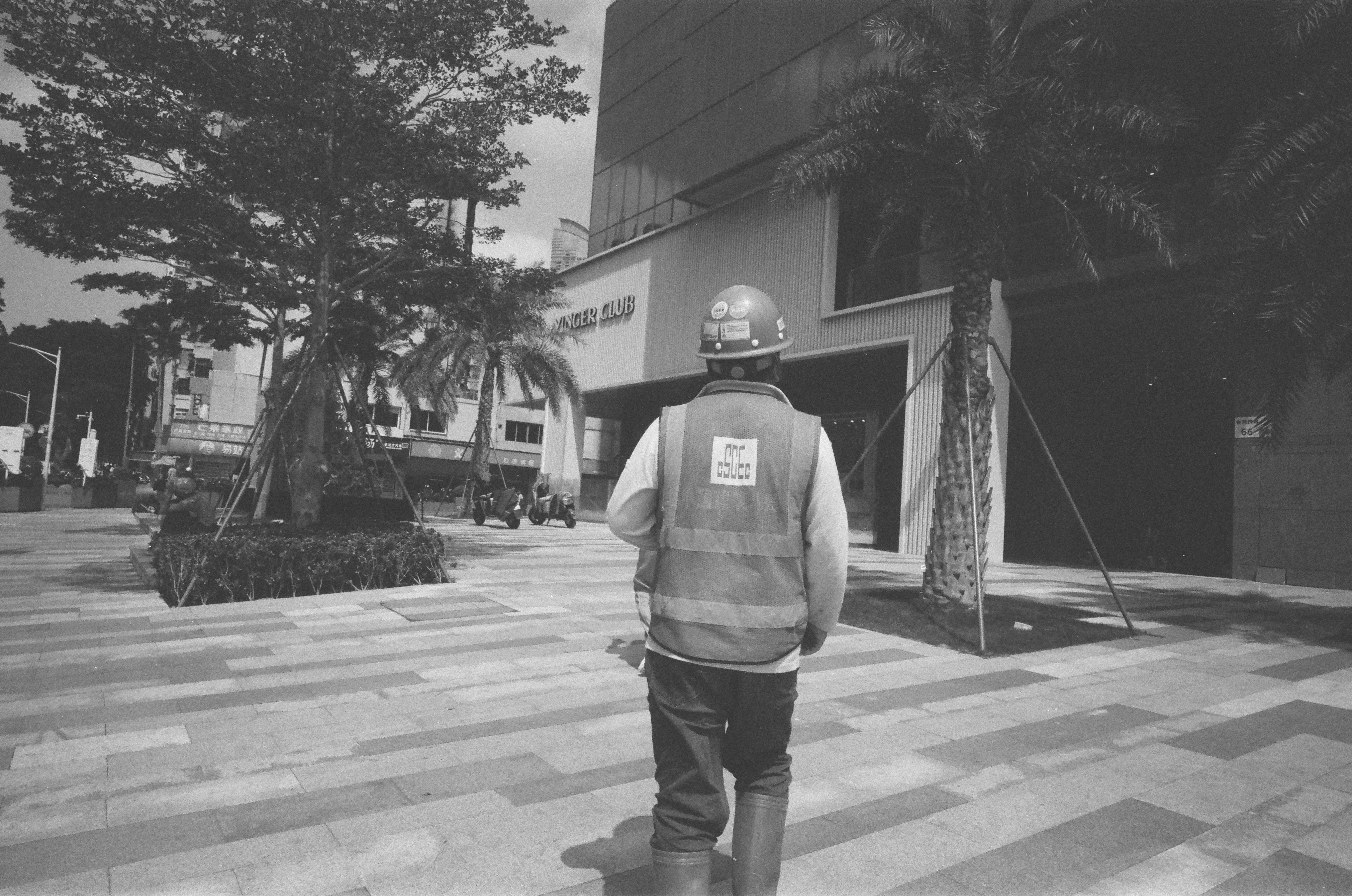 Un ouvrier du bâtiment en gilet de sécurité marche sur un trottoir à motifs.