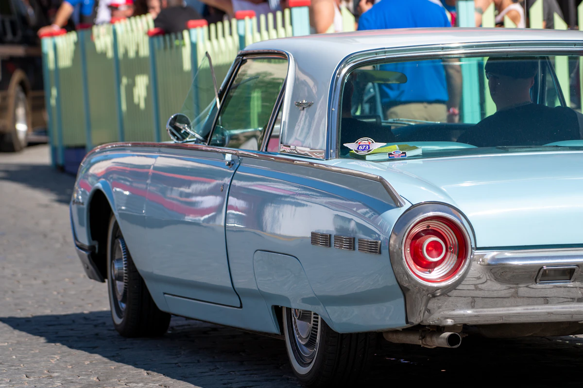 Light-blue classic Ford Thunderbird driving on a cobblestone street at a car show