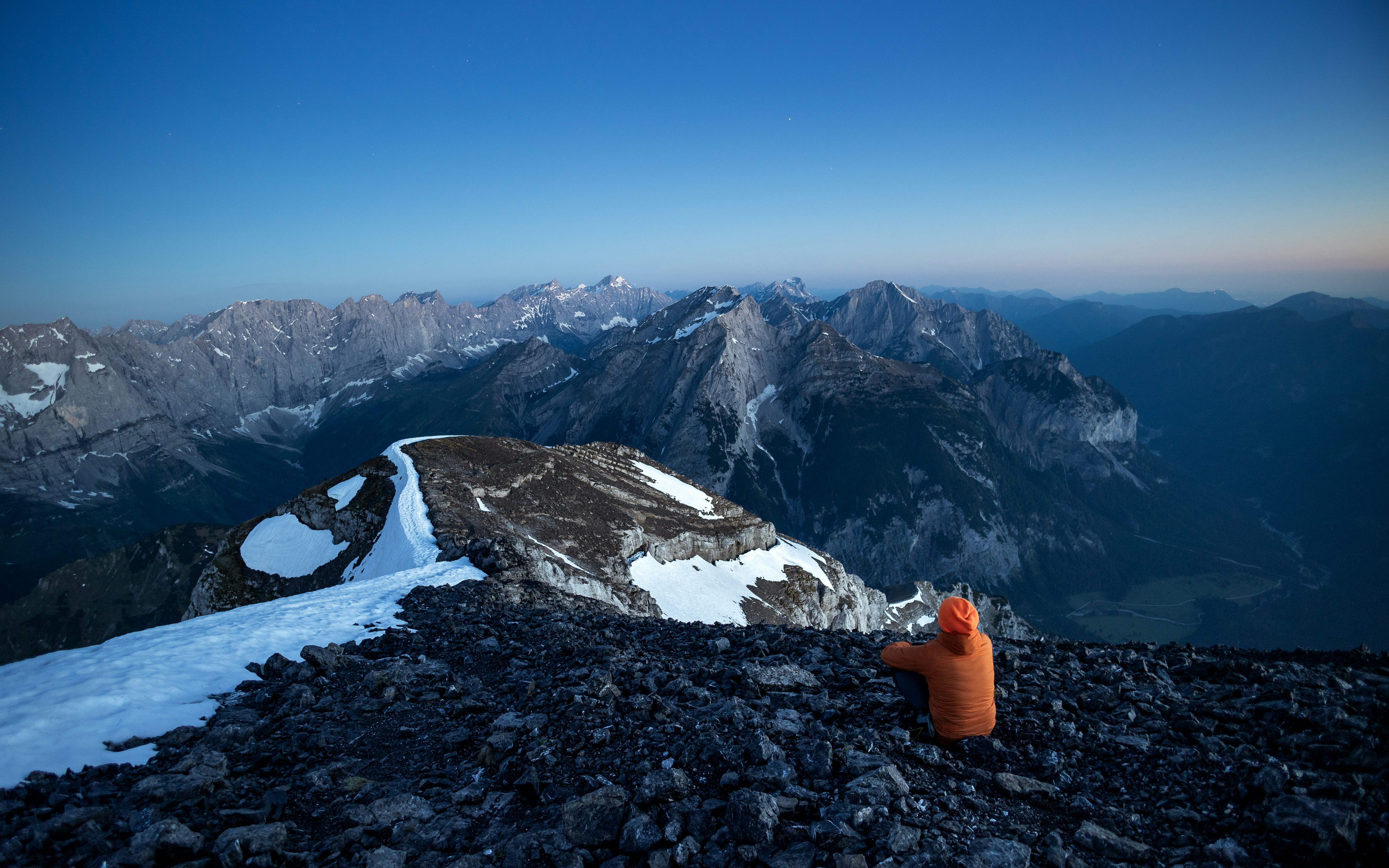 Person on rocky peak overlooks vast mountain range at dawn.