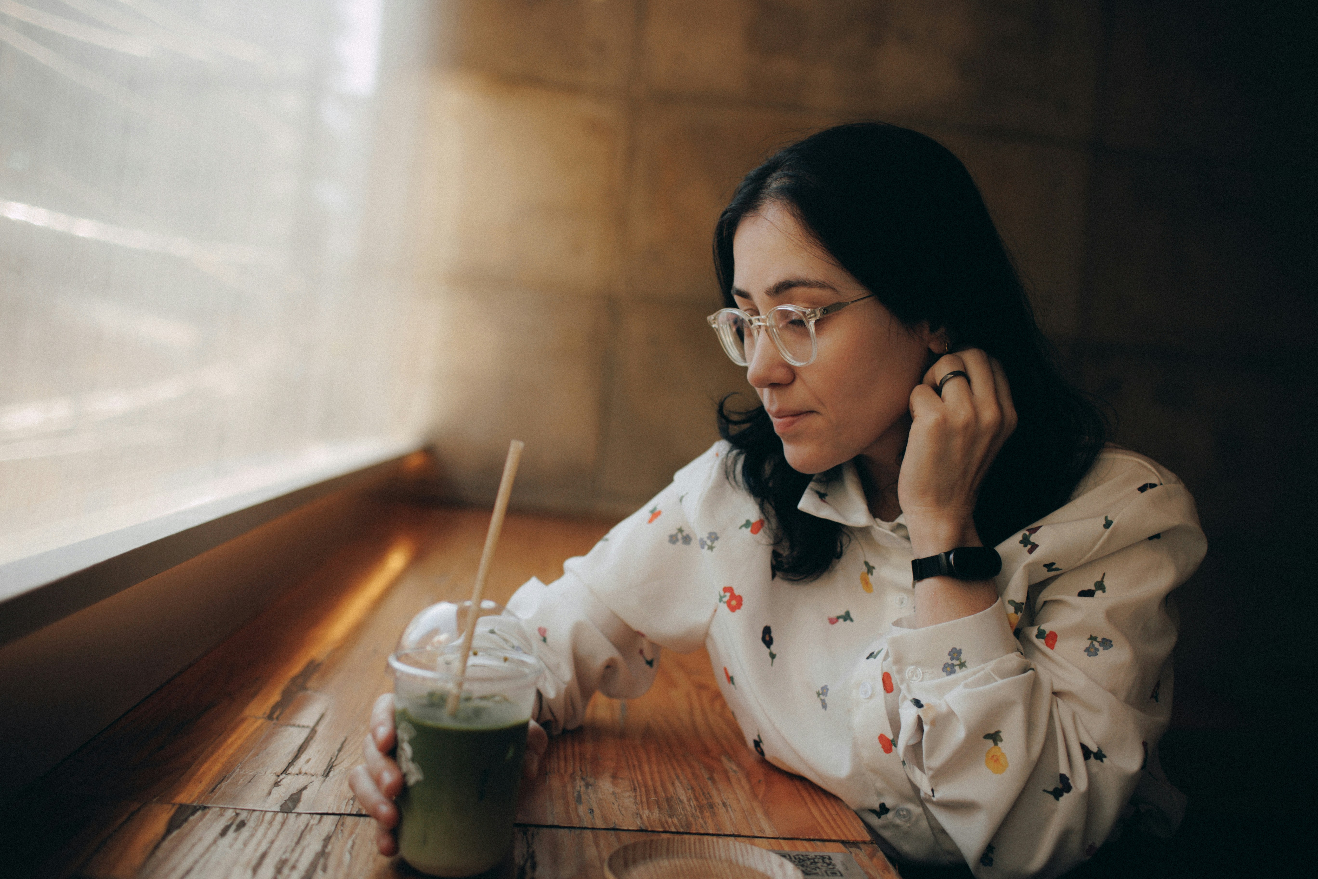 A contemplative woman with a green drink by a window.