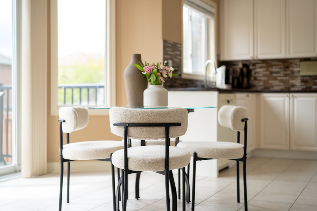 Small round glass dining table with white bouclé chairs and a vase of pink flowers in a bright apartment adjacent to the kitchen