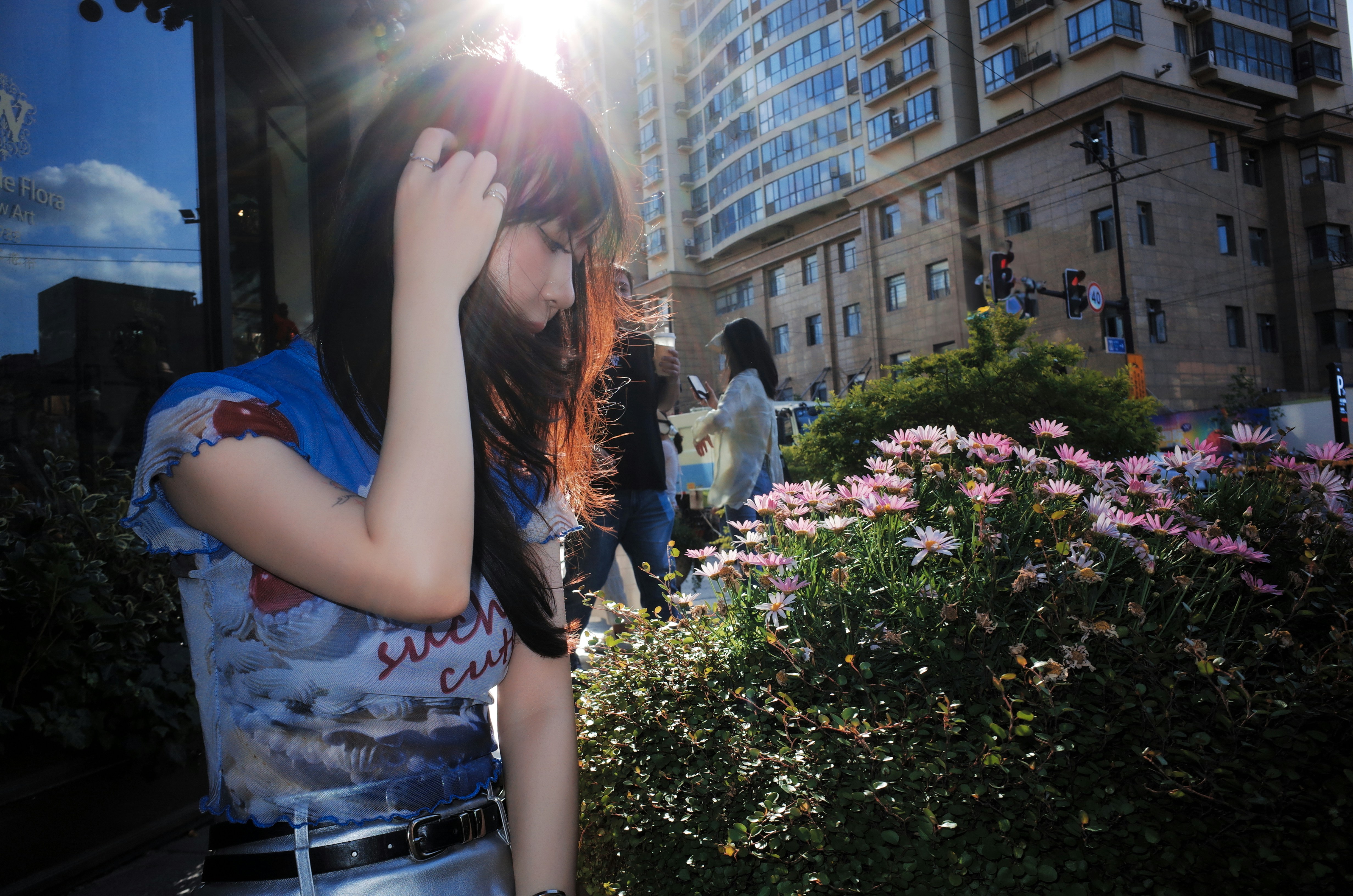 A young woman with sunlit hair stands among pink flowers.