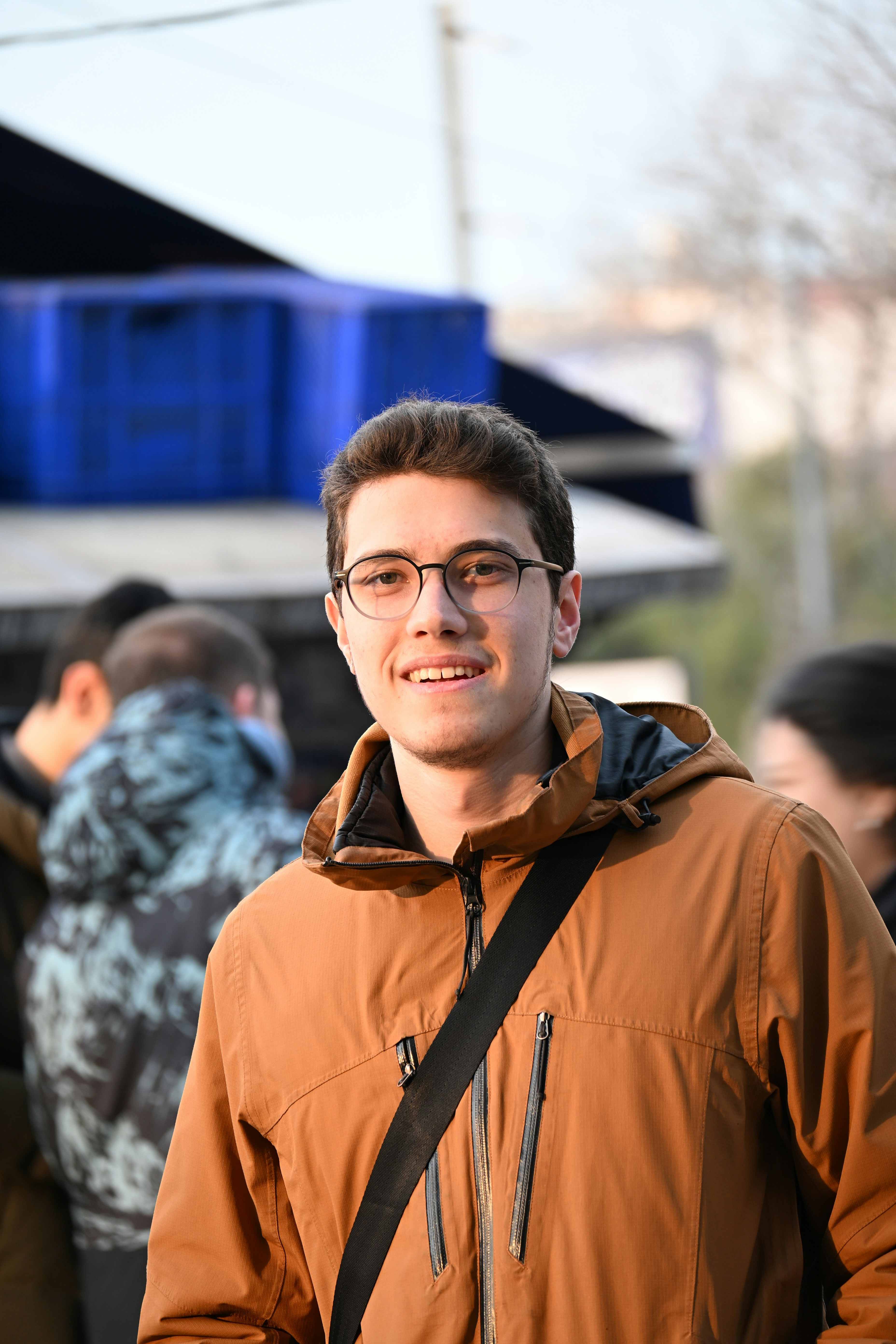 Un jeune homme à lunettes souriant dans une veste marron.