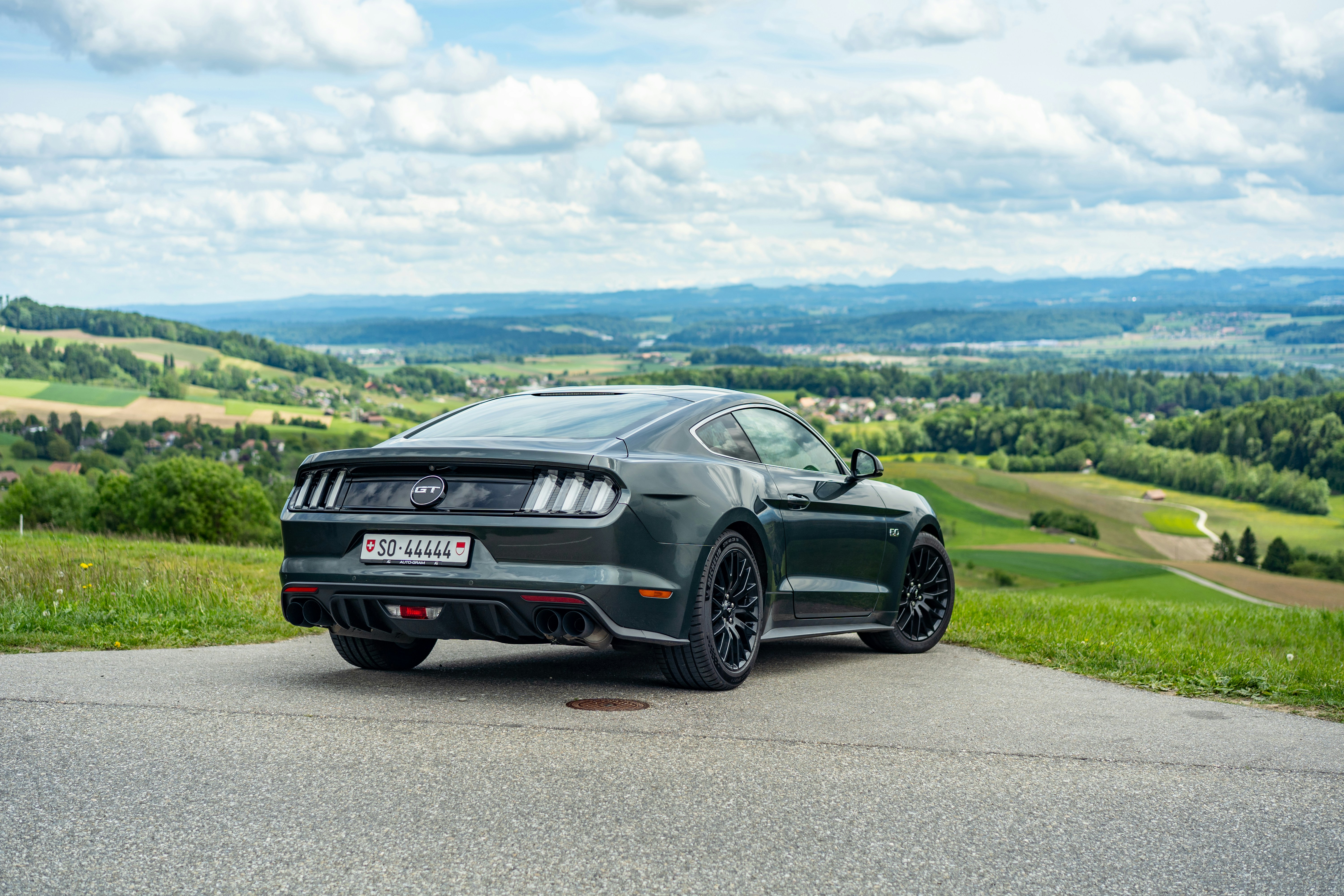 Voiture de sport sombre sur une route surplombant des collines verdoyantes.