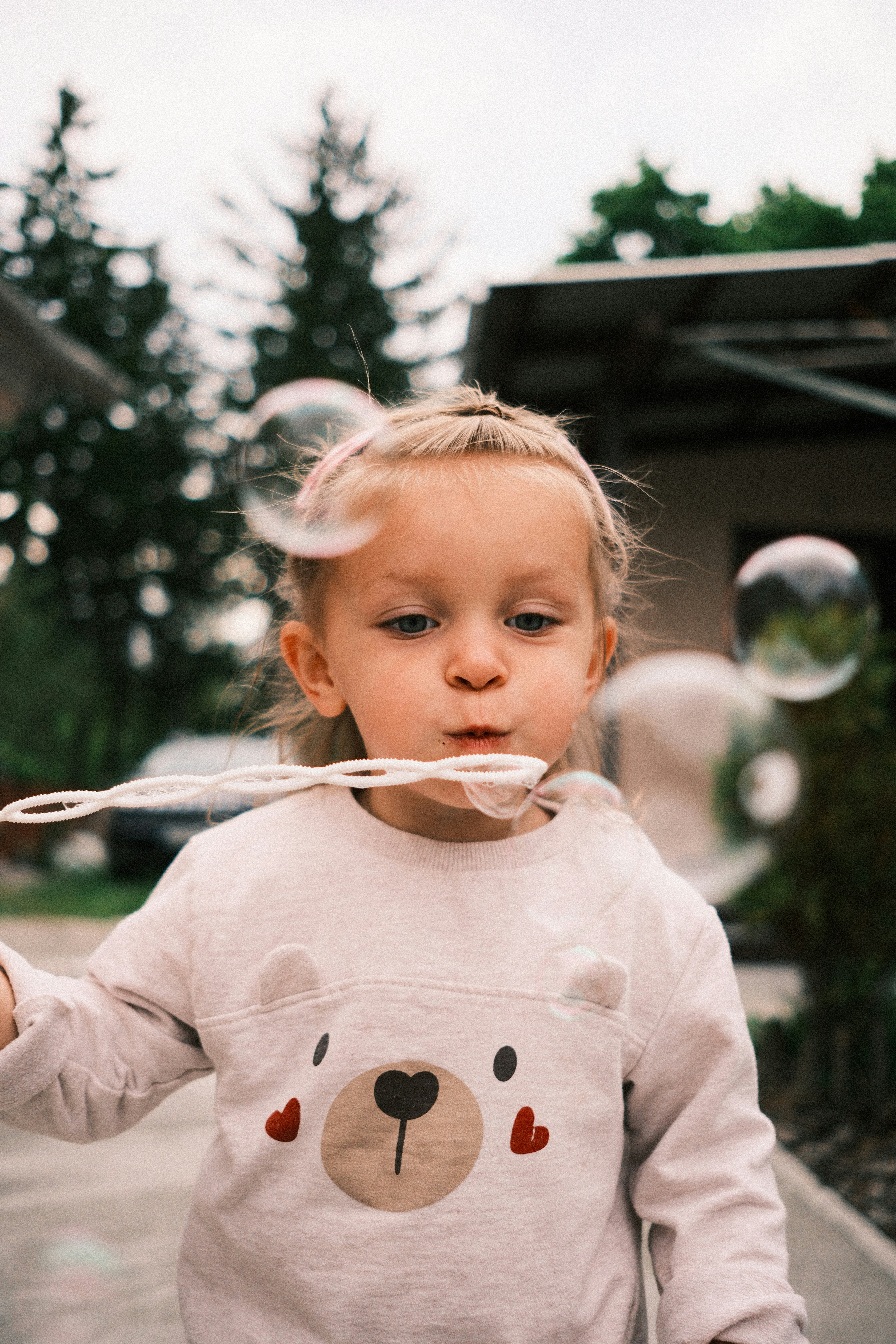 A young girl with blonde hair blows bubbles outdoors.