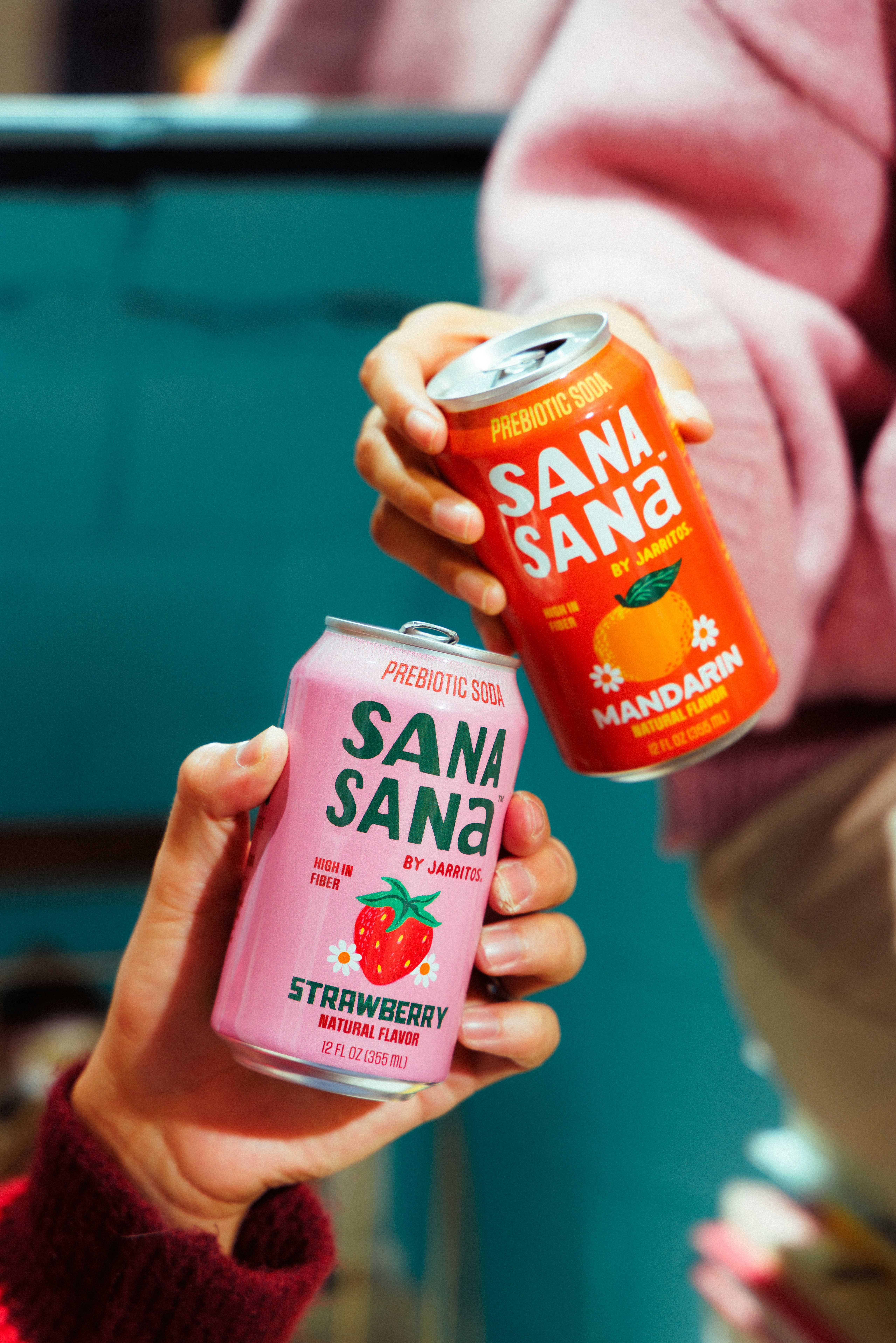 Dos manos sujetando latas de refresco sana sana de fresa y mandarina.